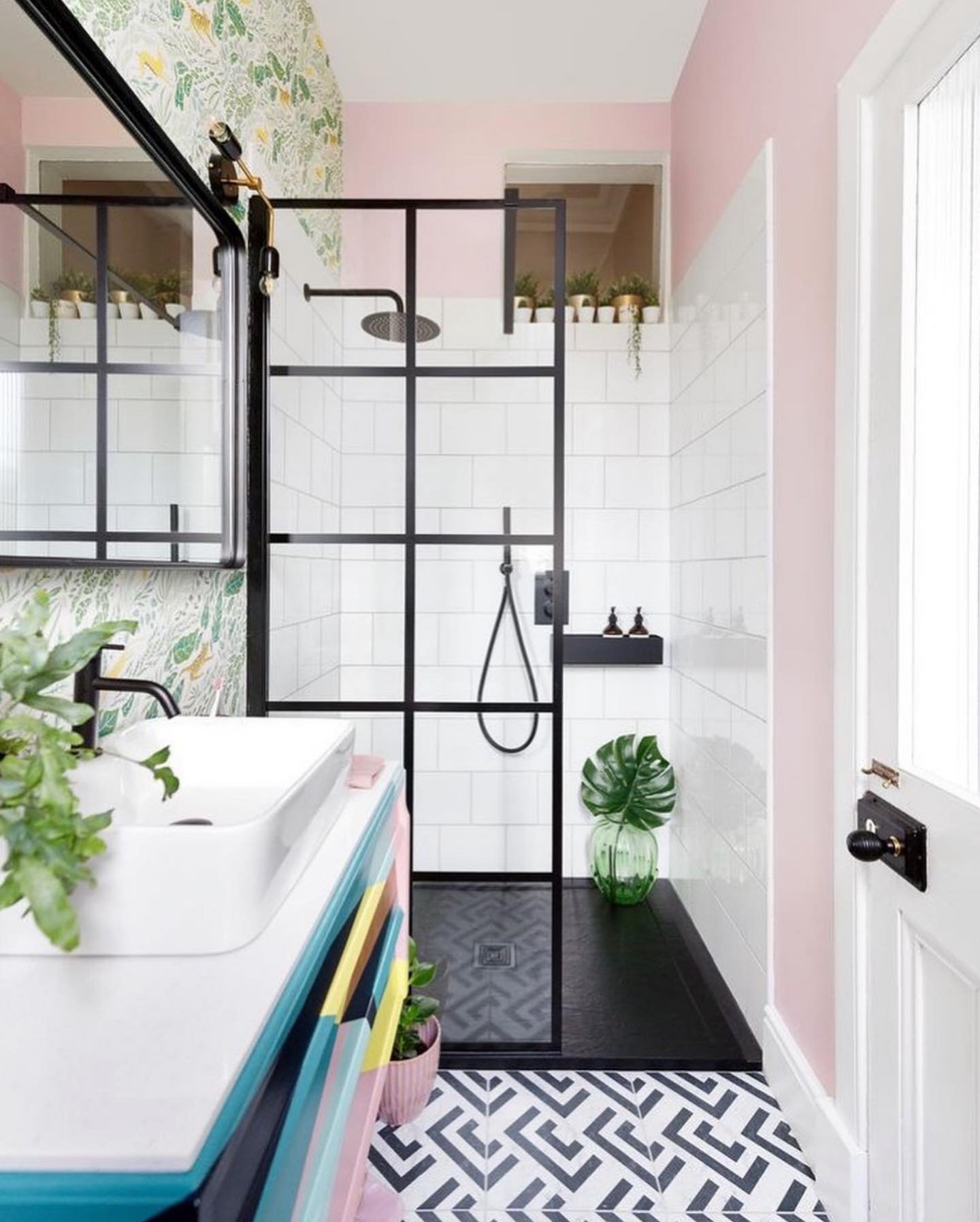 A vibrant small bathroom with a black and white geometric tiled floor, a pastel pink and plant-patterned wallpaper, and a shower with a black-framed glass enclosure.