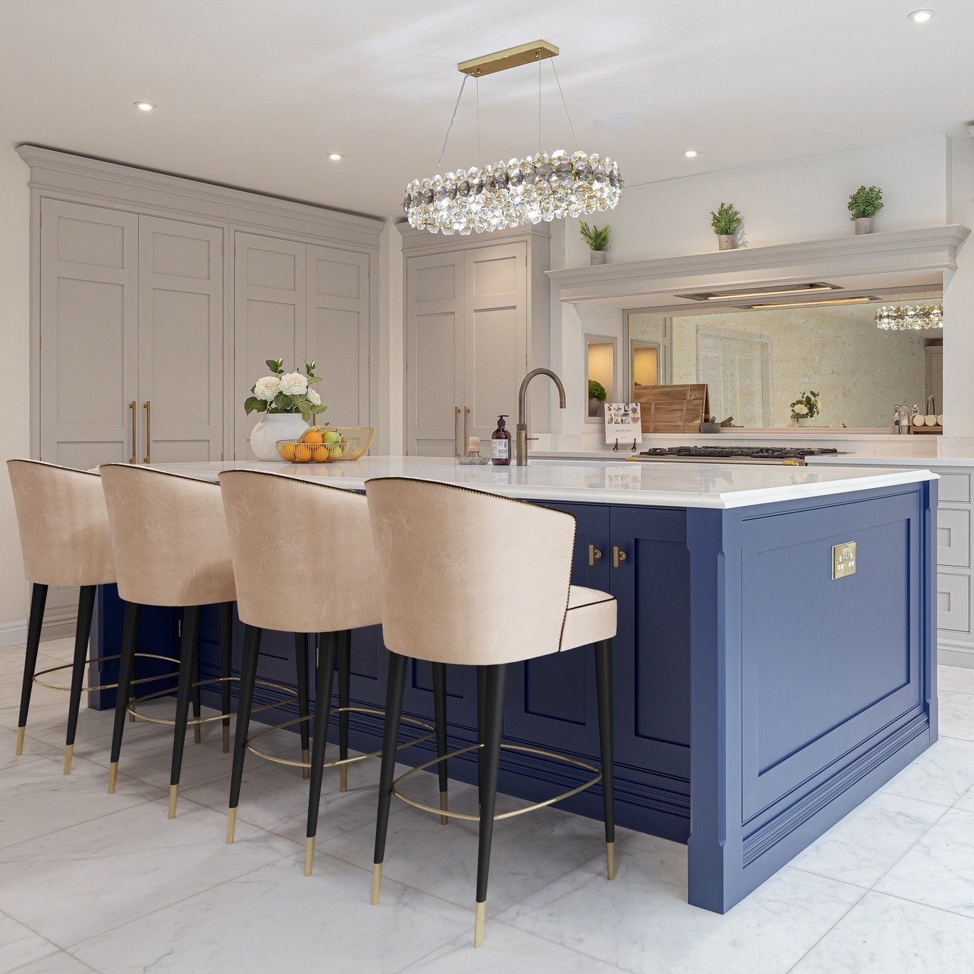 A large navy blue kitchen island with a white countertop, four beige velvet bar stools, and a crystal chandelier in a luxurious kitchen.