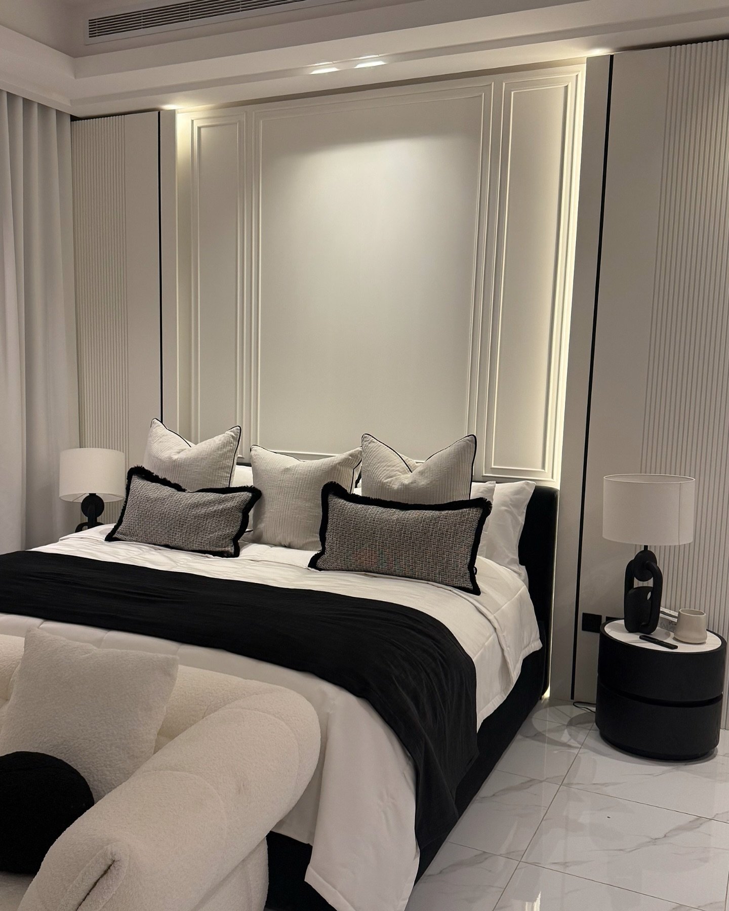 A sophisticated and luxurious guest bedroom with a black and white color scheme, featuring a king-sized bed, a minimalist paneled accent wall with integrated lighting, and a black circular nightstand.
