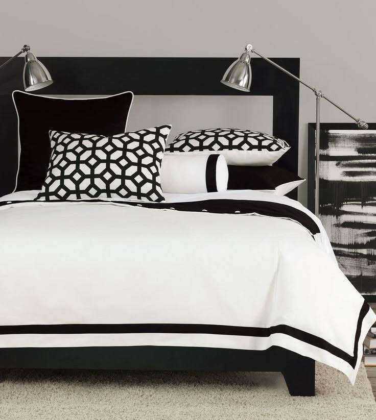 A stylish black and white bedroom with graphic geometric pillows, a clean white duvet with a black border, and two industrial-style floor lamps.