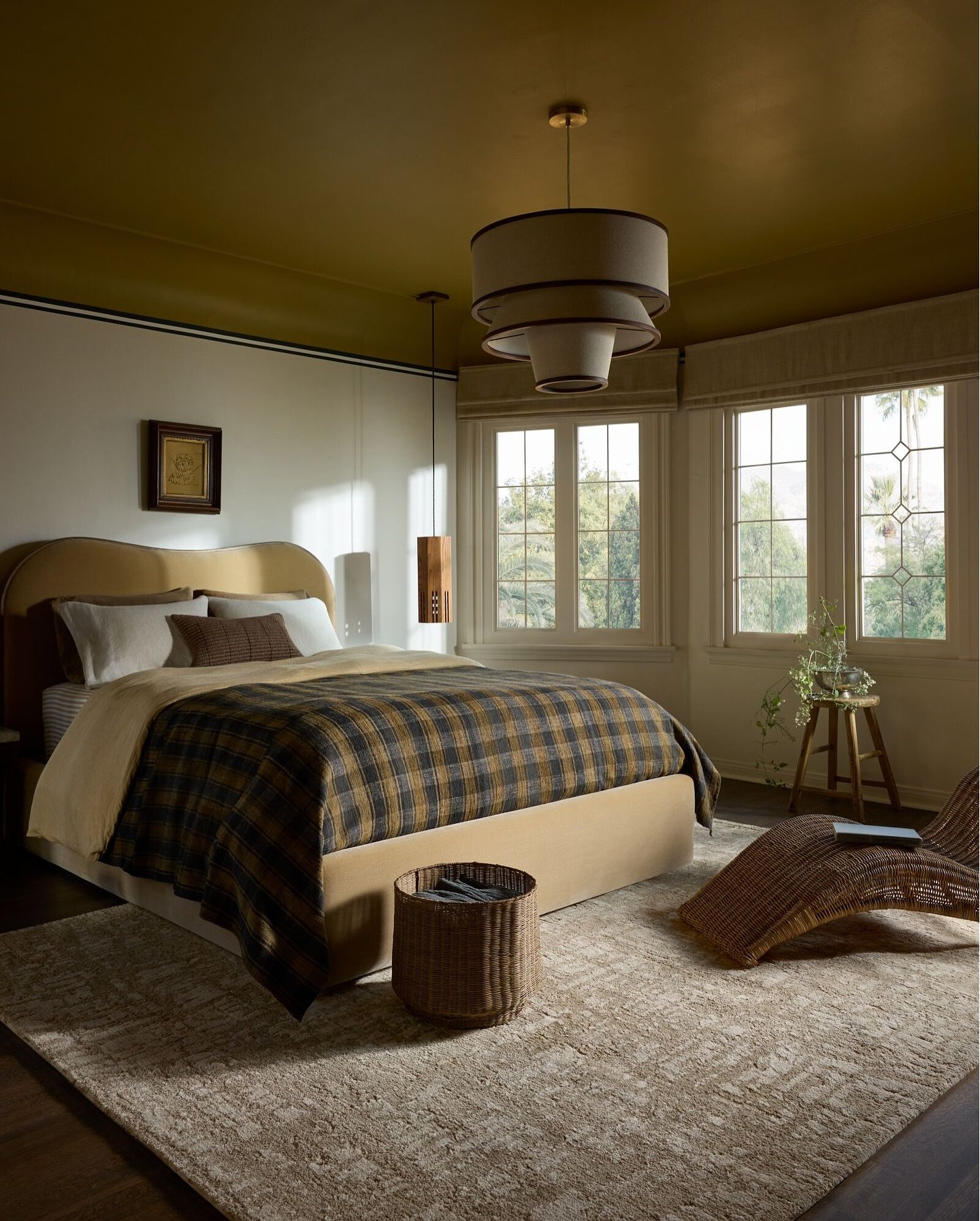 A large, light-colored area rug with a subtle plaid pattern in beige and brown is placed under a bed with a dark plaid blanket.