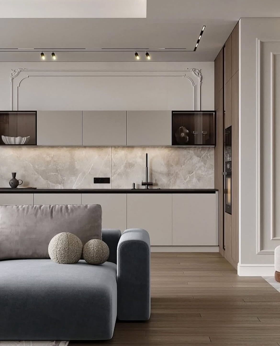 A close-up view of a modern open-concept space. In the foreground is a dark grey sectional sofa. The kitchen features a mix of flat grey cabinets and glass-front cabinets, with a striking marble backsplash and a contrasting panel of white molding and wood on the side.