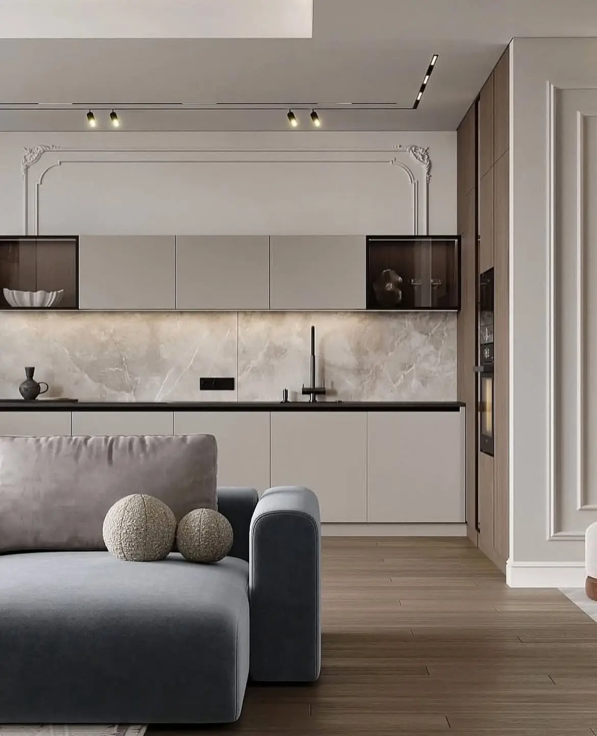 A close-up view of a modern open-concept space. In the foreground is a dark grey sectional sofa. The kitchen features a mix of flat grey cabinets and glass-front cabinets, with a striking marble backsplash and a contrasting panel of white molding and wood on the side.