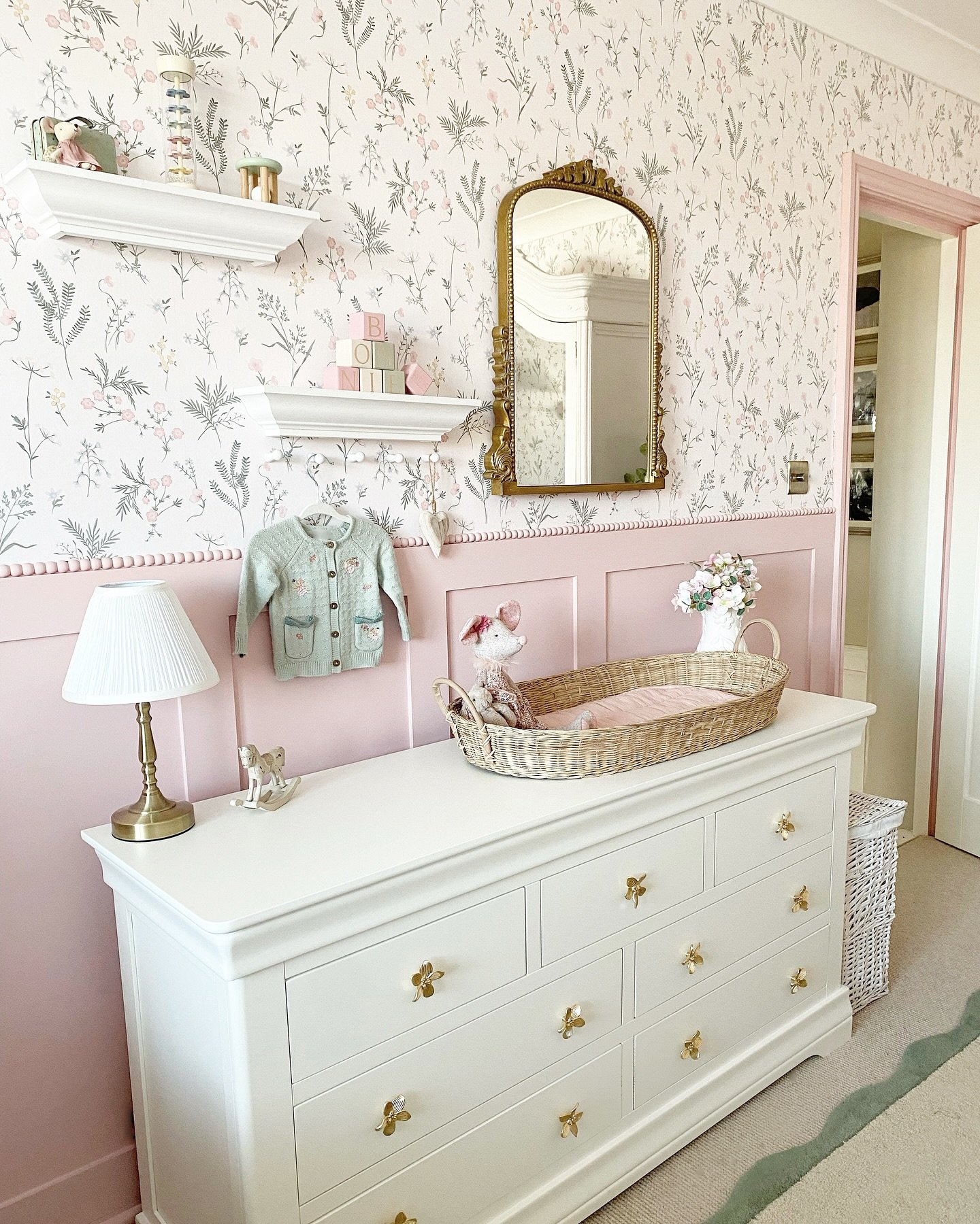An elegant nursery with a pink and floral wall, a white dresser with butterfly knobs, and a gold mirror.