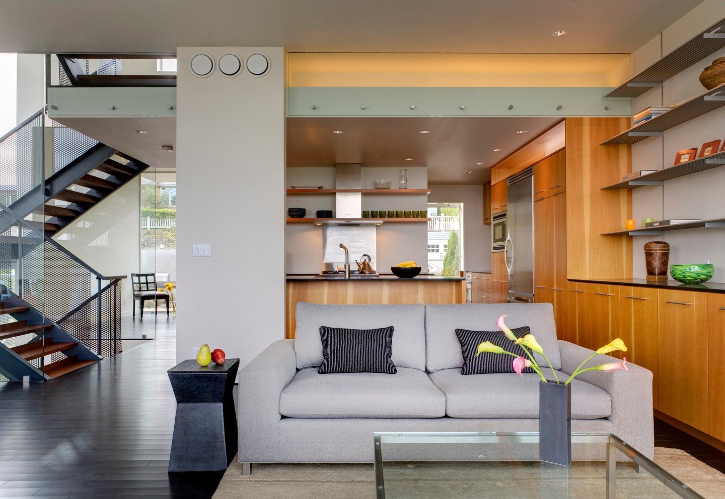 A view of a modern, multi-level home with an open-concept kitchen and living room. A light grey sofa and glass coffee table are in the foreground. The kitchen features light wood cabinetry and a peninsula. A metal staircase with wood treads leads to an upper floor on the left.