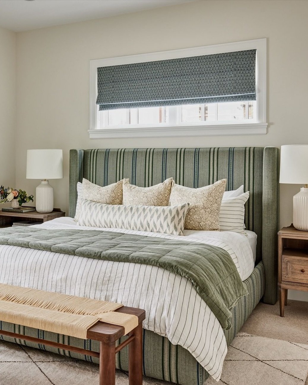 A stylish guest bedroom with a striped upholstered bed and a variety of layered patterns and textures. The room features a woven bench at the foot of the bed, ribbed lamps, and rustic wood nightstands.