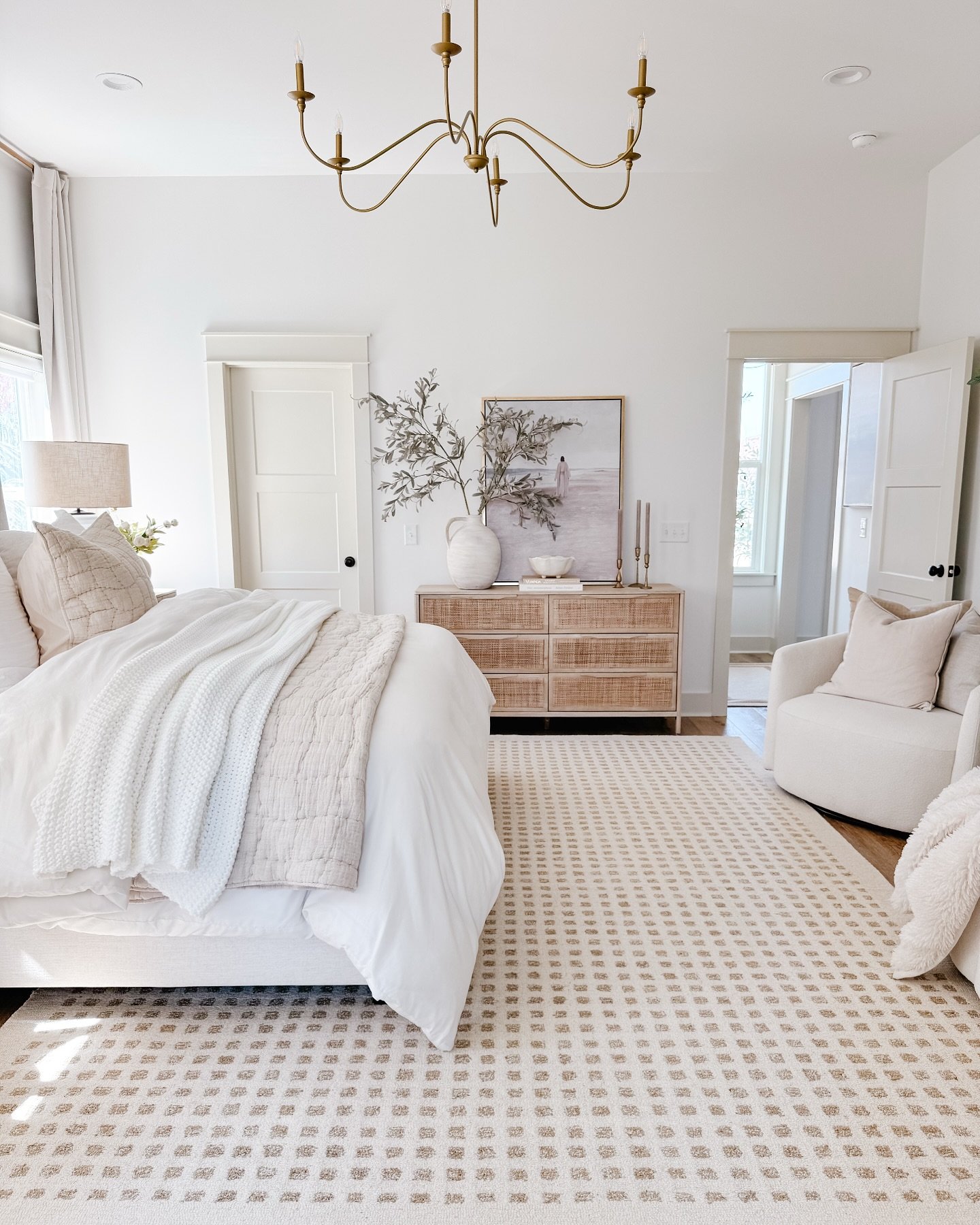 A large, light-colored area rug with a repeating square grid pattern in beige and cream is placed in a bright, white bedroom.