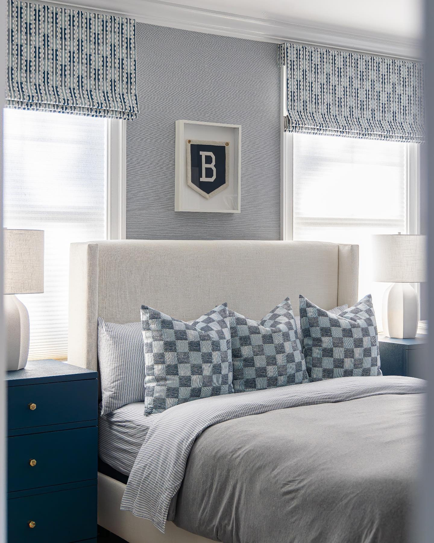 A classic teen's bedroom with a navy blue and gray color scheme, featuring a textured wallpaper, roman blinds, and an initial pennant.