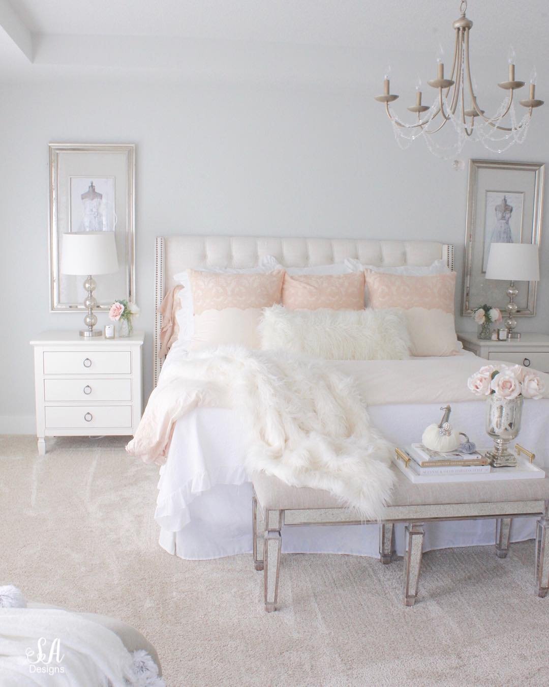 A glamorous bedroom with a tufted headboard, soft pink and white bedding, and a mirrored bench at the foot of the bed.