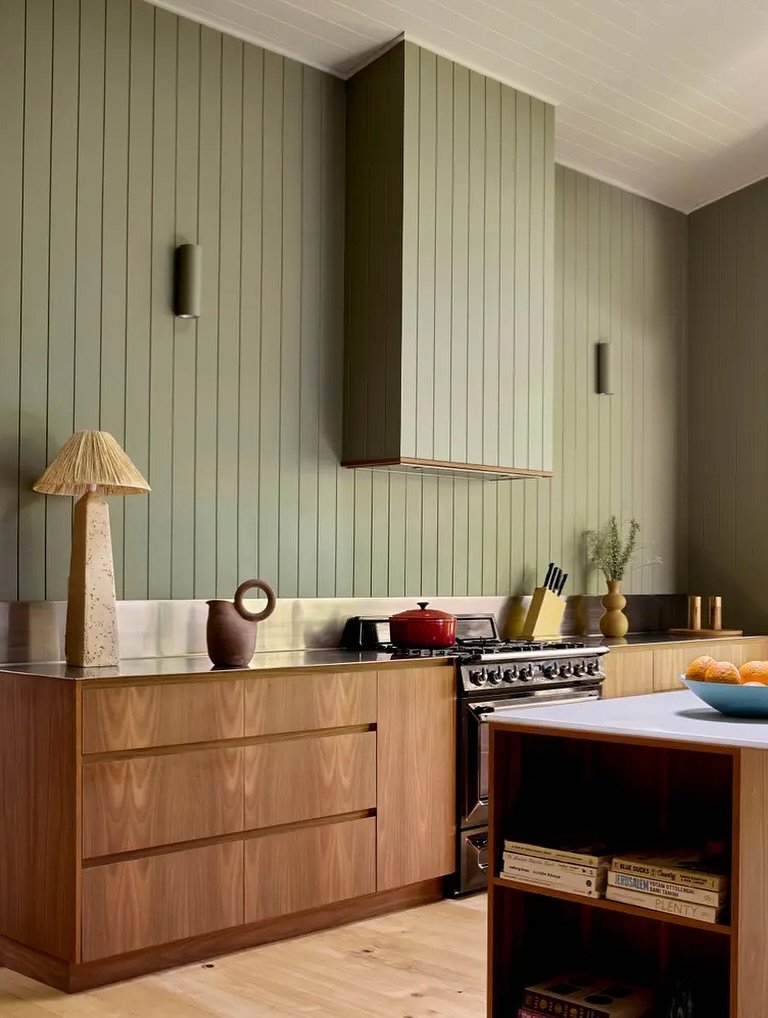 A contemporary kitchen with a low, seamless stainless steel backsplash, set against a backdrop of sage green vertical plank walls and warm wood cabinetry.