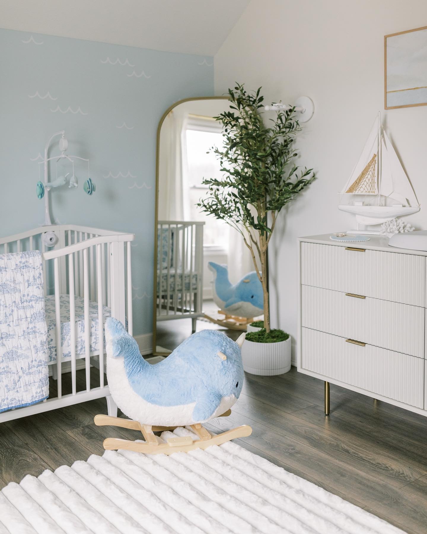 A serene nursery with a light blue wall, a white crib, a large whale rocking toy, and a gold-framed mirror.