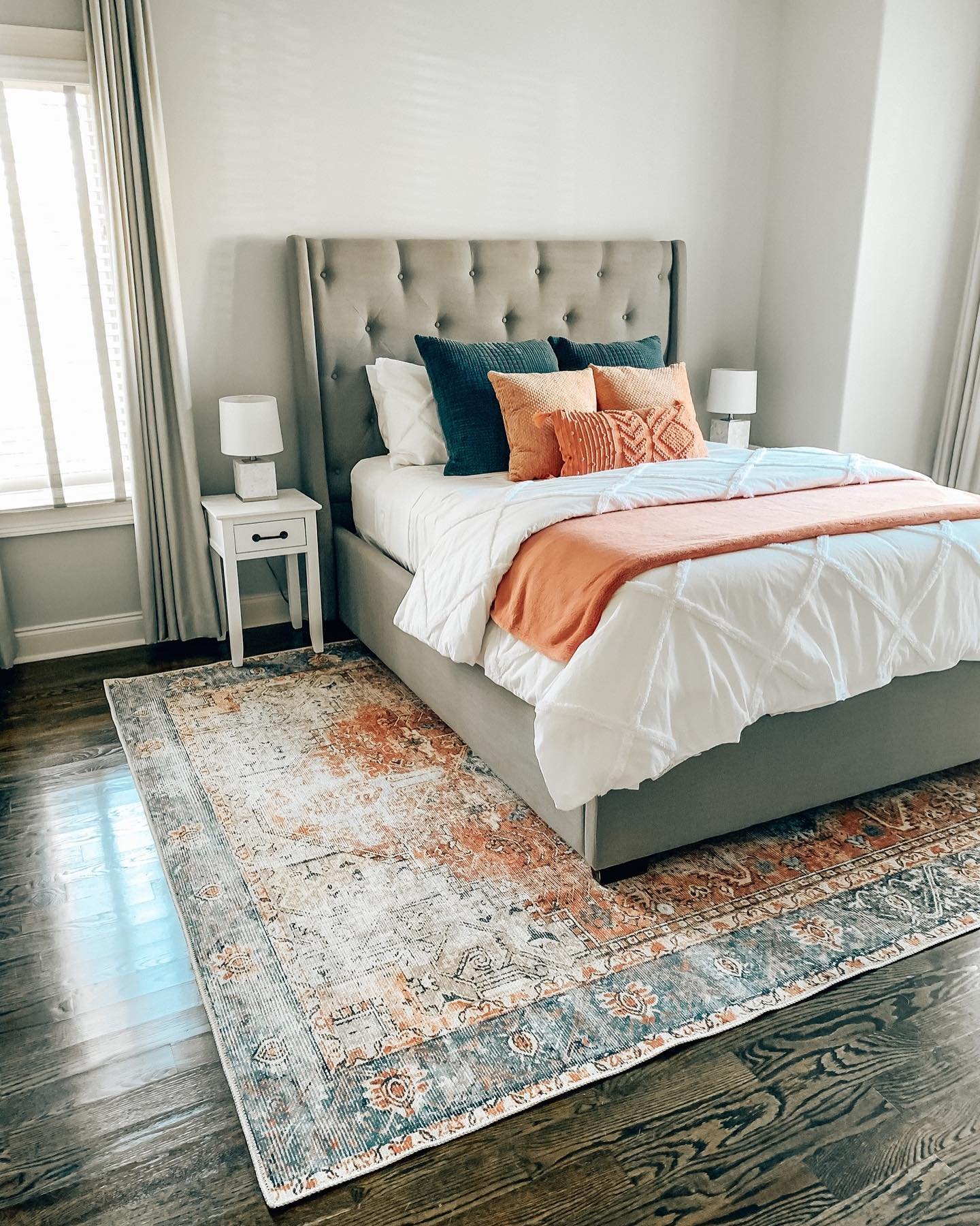 A modern guest bedroom with a gray tufted headboard, a white comforter, and bright terracotta and navy accent pillows and a throw. A distressed orange and blue patterned rug covers the dark wood floor.