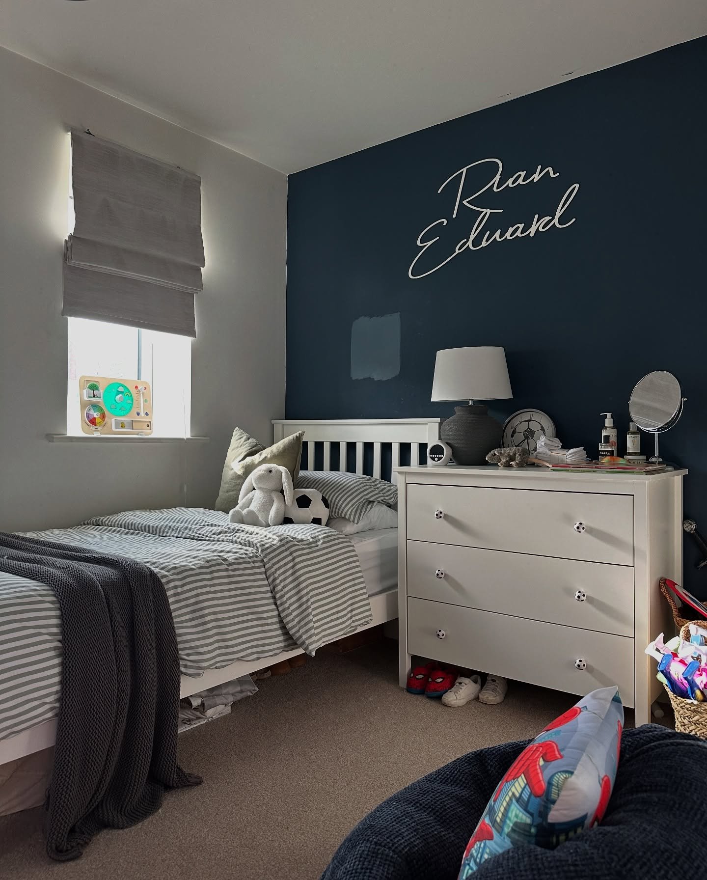 A teenage bedroom with a white bed, a dark blue accent wall with a custom name sign, and a white dresser.