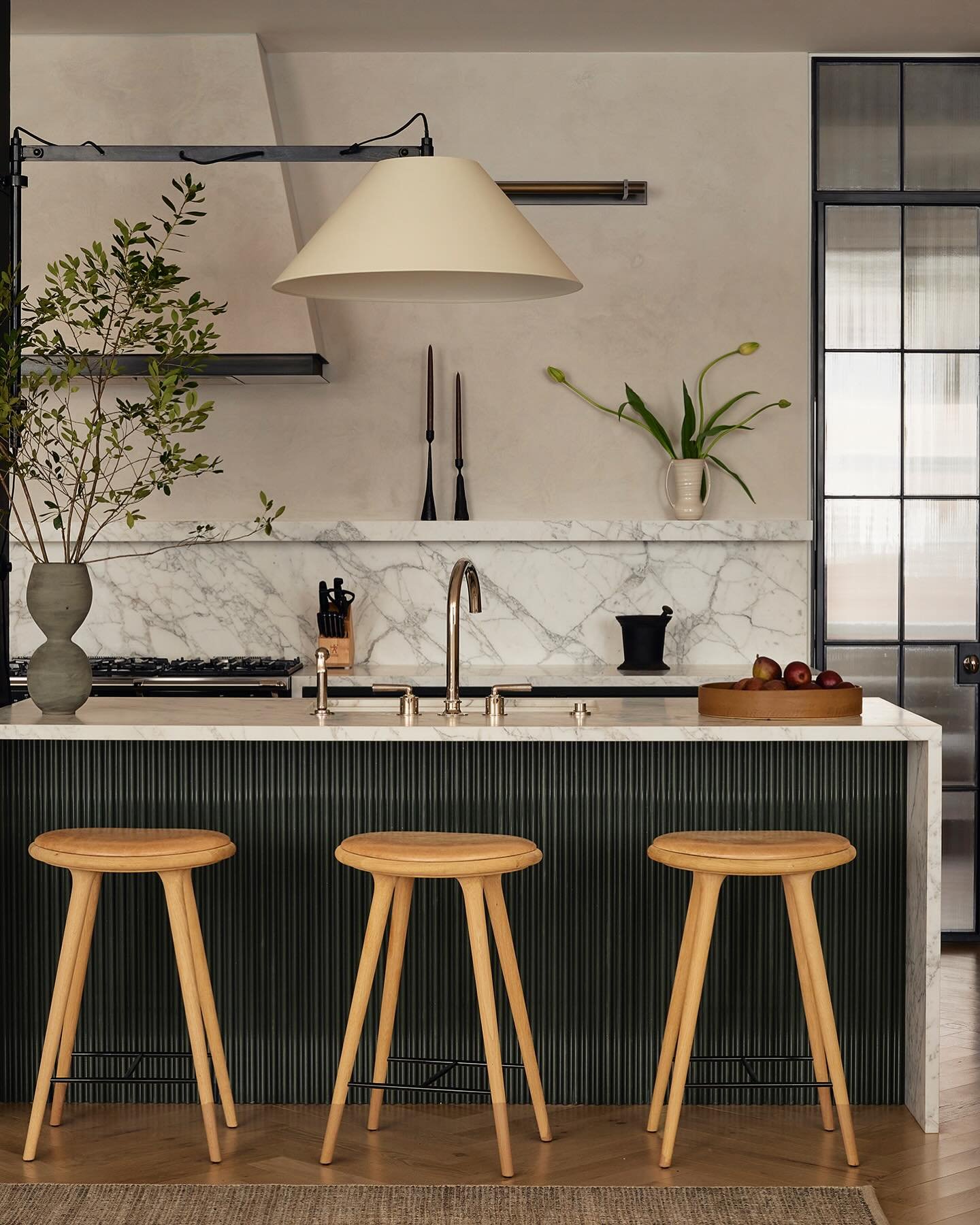 A small kitchen island with a black fluted base, a marble countertop, and three light wooden bar stools in a stylish kitchen.