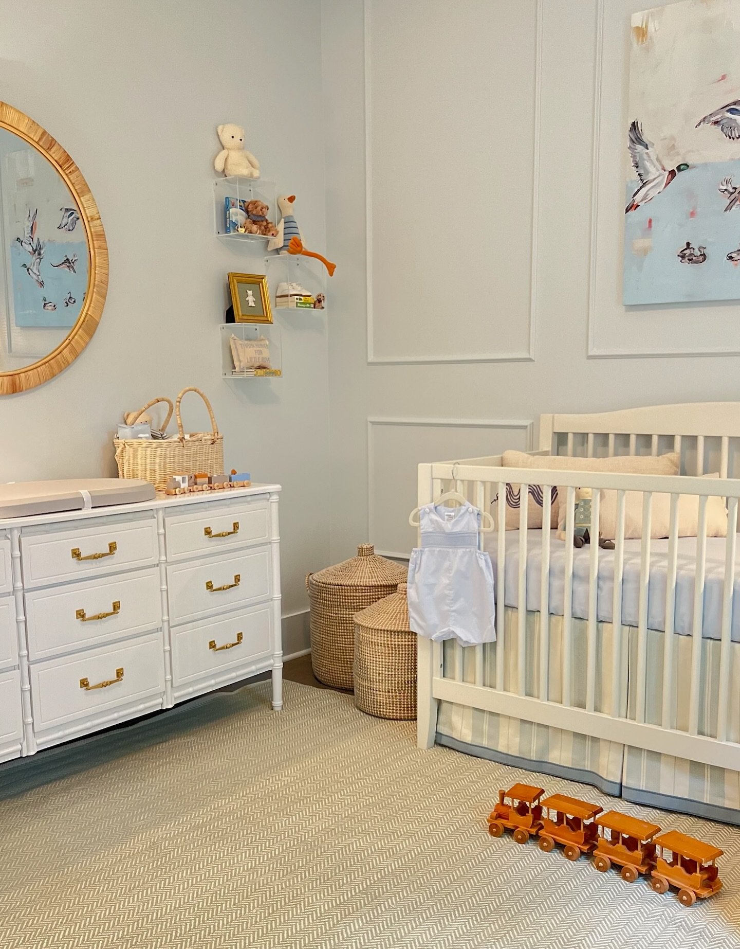 A traditional light blue nursery room with a white dresser, round rattan mirror, and a white crib.