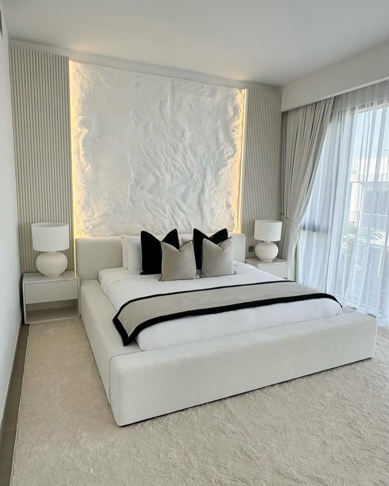  A luxurious all-white bedroom with a textured and illuminated headboard wall, a low-profile plush bed, and matching ribbed nightstands with minimalist lamps.