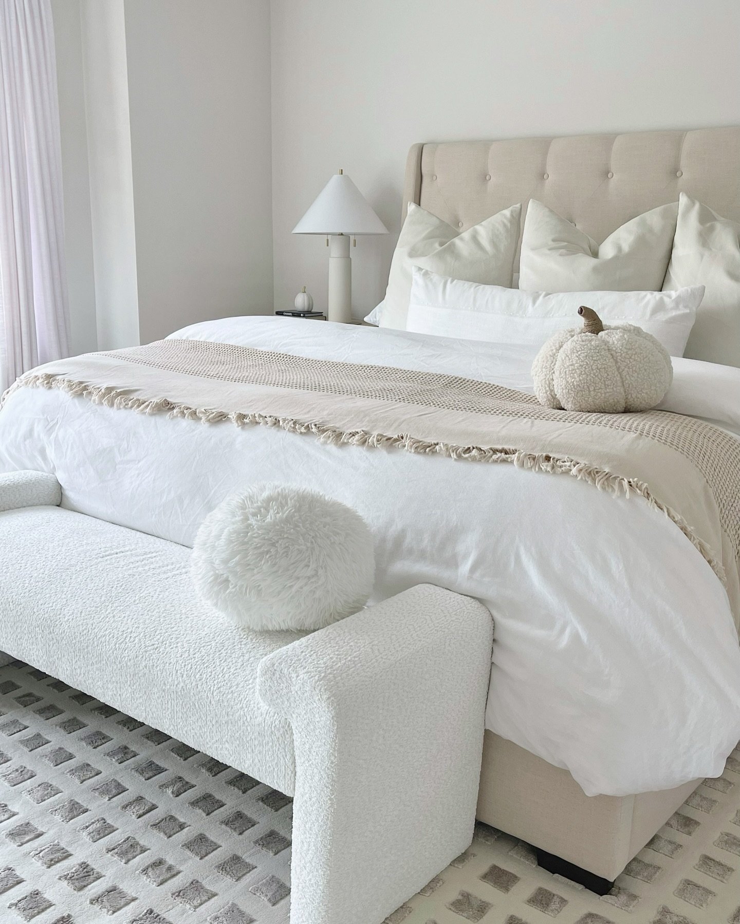A bright, minimalist bedroom featuring a bed with a tufted headboard and white and beige layered bedding. A white bouclé bench with a furry round pillow sits at the foot of the bed, next to a textured, cream-colored pumpkin accent pillow.