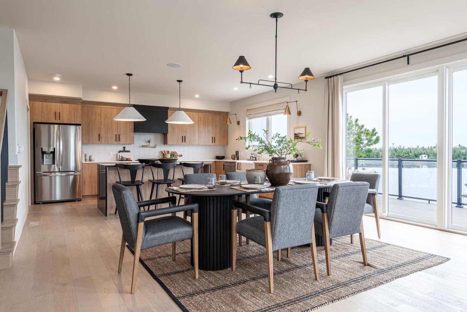 A contemporary open-concept kitchen and dining area with a coastal feel. A large round dining table with grey upholstered chairs sits on a textured rug. In the background is a kitchen with light wood cabinets, a black range hood, and a large island with stools. Sliding glass doors open to a water view.