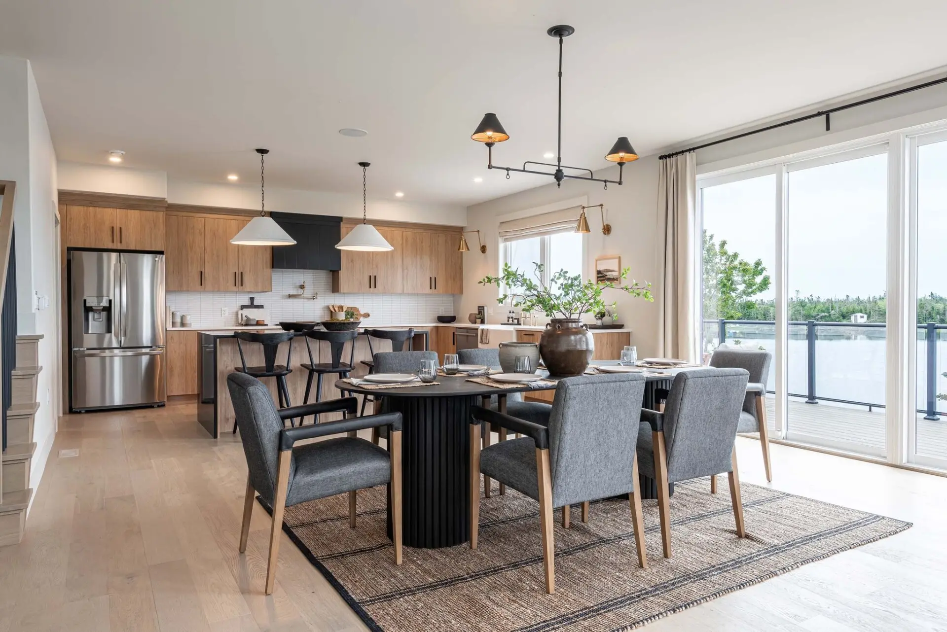 A contemporary open-concept kitchen and dining area with a coastal feel. A large round dining table with grey upholstered chairs sits on a textured rug. In the background is a kitchen with light wood cabinets, a black range hood, and a large island with stools. Sliding glass doors open to a water view.