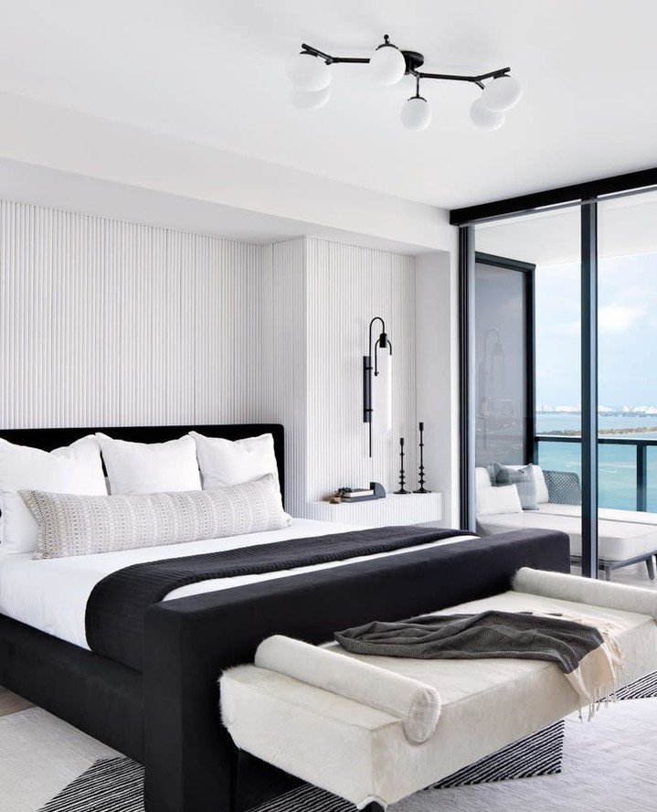 A luxurious modern bedroom with a black upholstered bed, a white ribbed accent wall, and a large sliding glass door with a balcony overlooking the ocean.