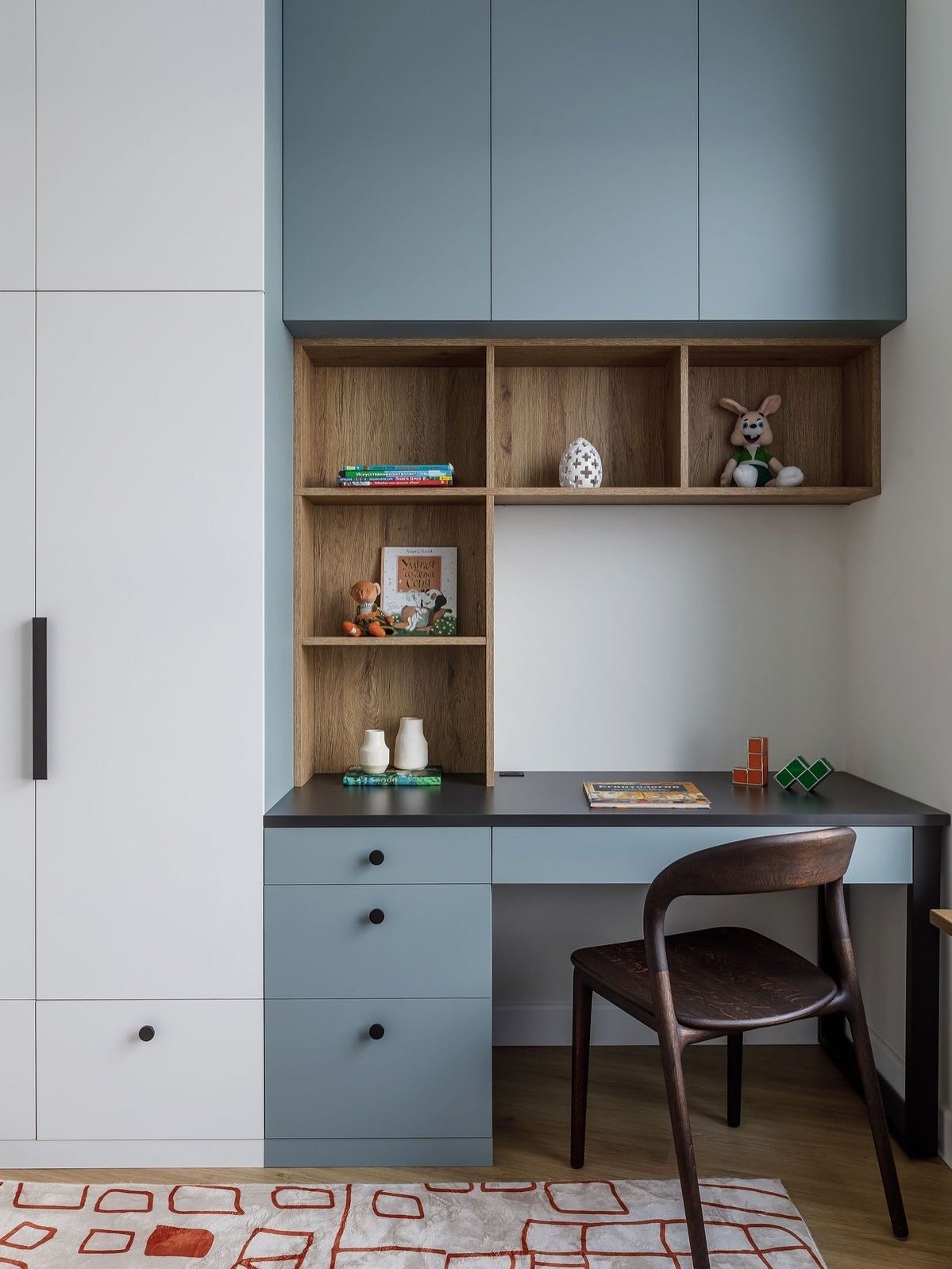 A compact, integrated study desk with a dark grey top and light blue-grey drawers, a wooden shelf unit above, and a dark wooden chair.