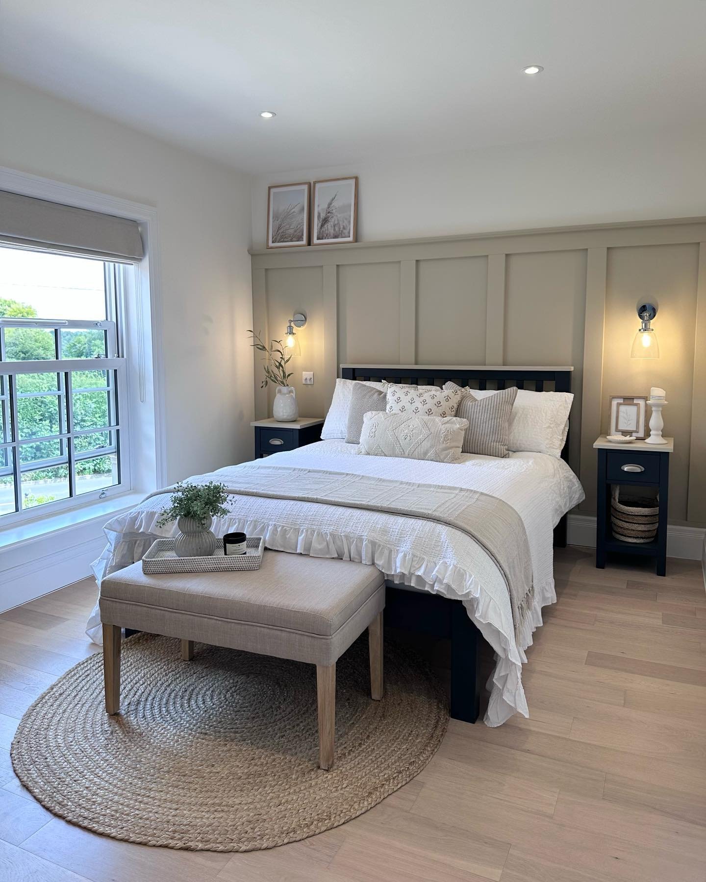 A cozy guest bedroom with a dark wood bed frame, light blue nightstands, and a board and batten accent wall. The room is decorated in a coastal farmhouse style with a jute rug, a neutral upholstered bench, and framed art.