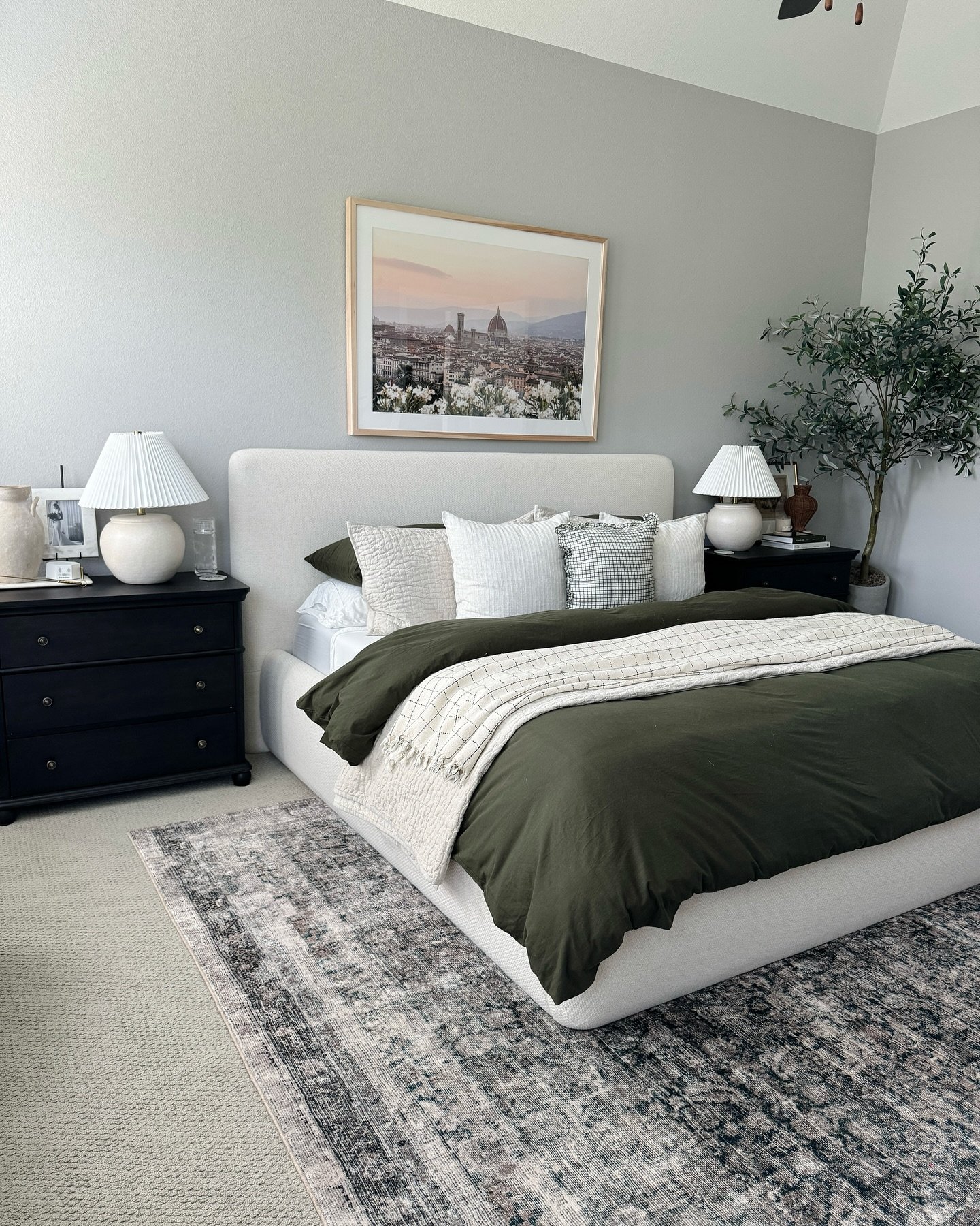 A bedroom with light gray walls, a white upholstered bed, and a large piece of art featuring a cityscape at sunset above the headboard. 
