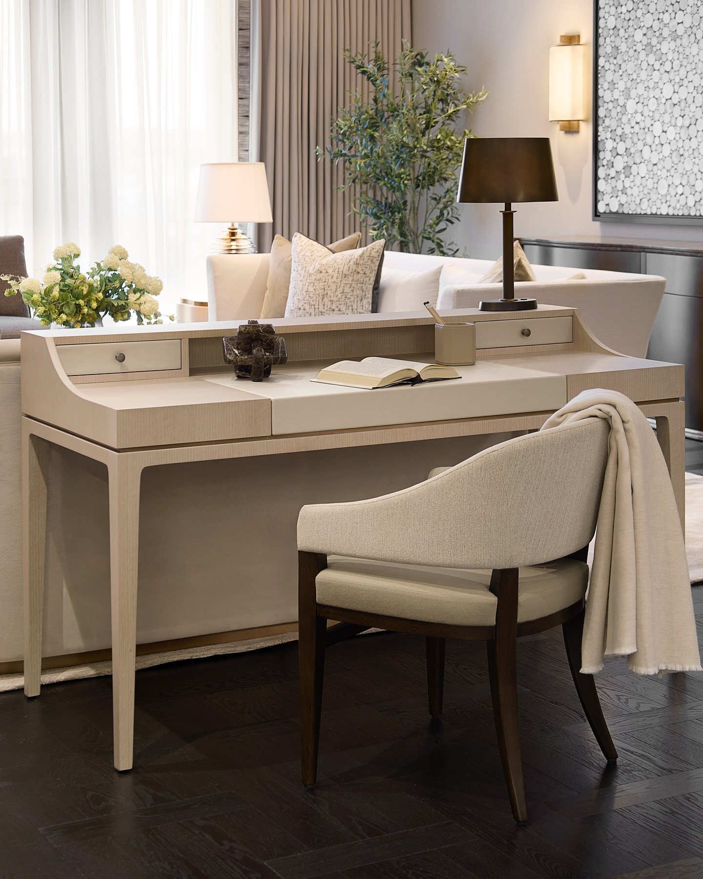 An elegant and minimalist light-toned wooden writing desk with small drawers, an upholstered chair, and a table lamp on the surface.