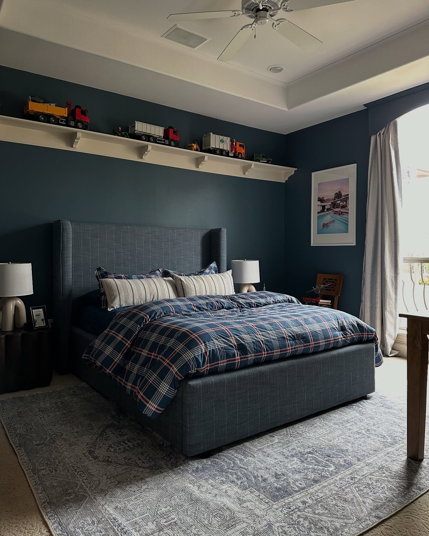A teenage boy's bedroom with dark blue walls, a bed with plaid and striped bedding, and a high shelf displaying toy trucks.