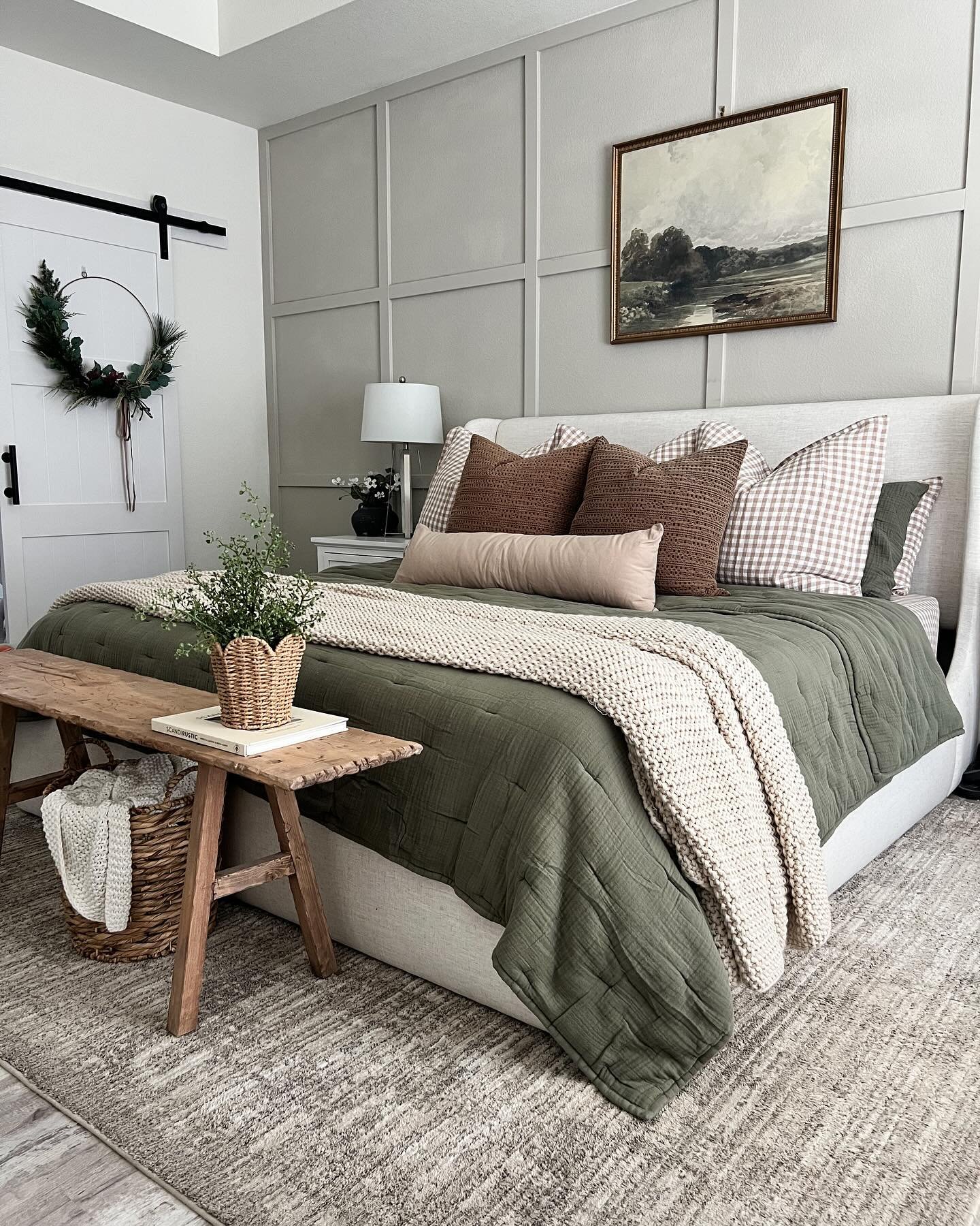 A cozy farmhouse-style guest bedroom with a queen-sized bed featuring green and terracotta bedding, a light gray wainscoting accent wall, and a rustic wood bench at the foot of the bed.