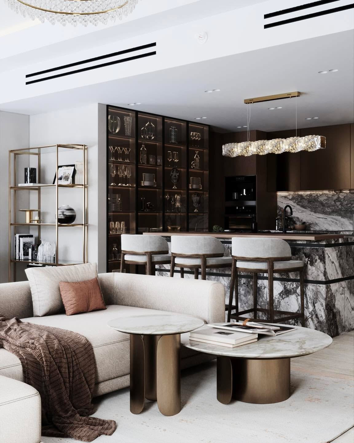 A high-end, open-concept living and kitchen area. The living room features a light-colored sectional sofa and two round marble-topped coffee tables. The kitchen has a stunning marble island with three bar stools, dark wood cabinetry, and a glass display case filled with decor.