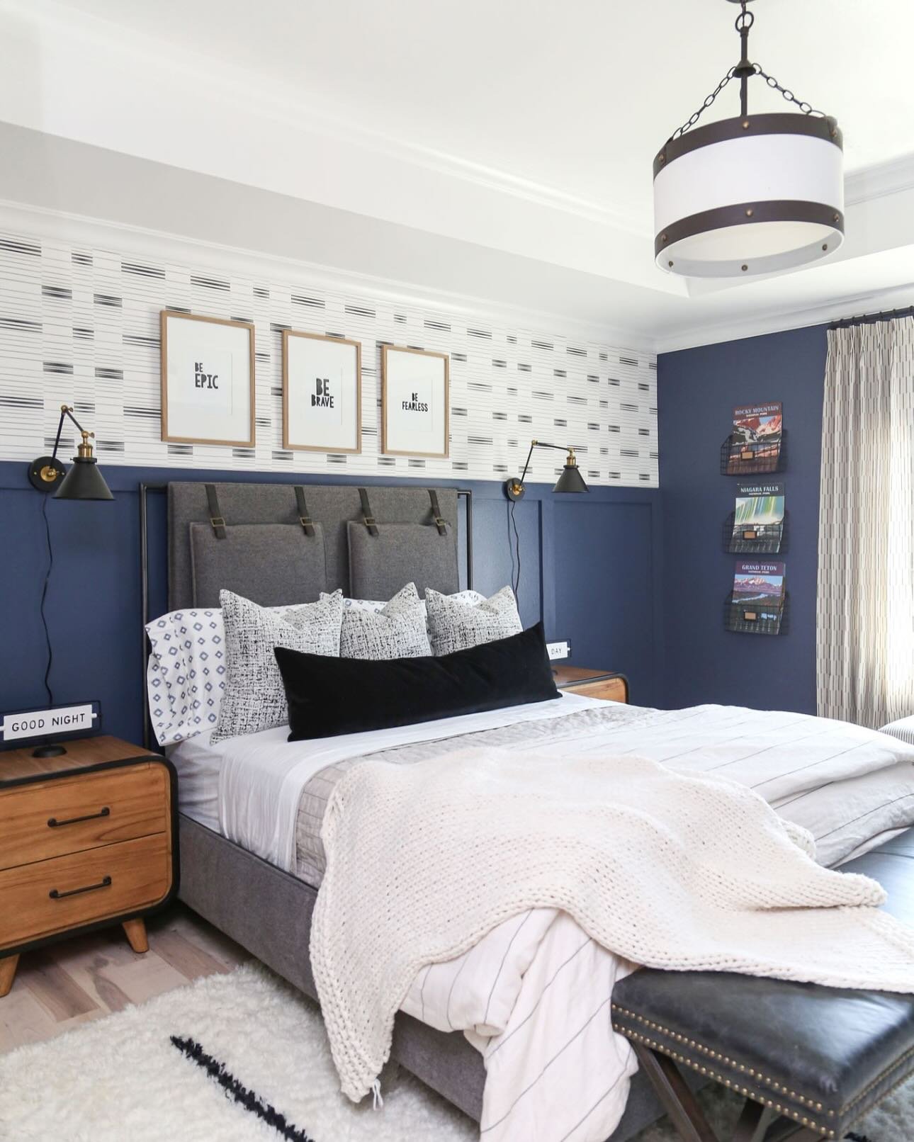 A teenage boy's bedroom with a two-toned wall—dark blue on the bottom and a graphic patterned wallpaper on top—and a bed with a gray upholstered headboard.
