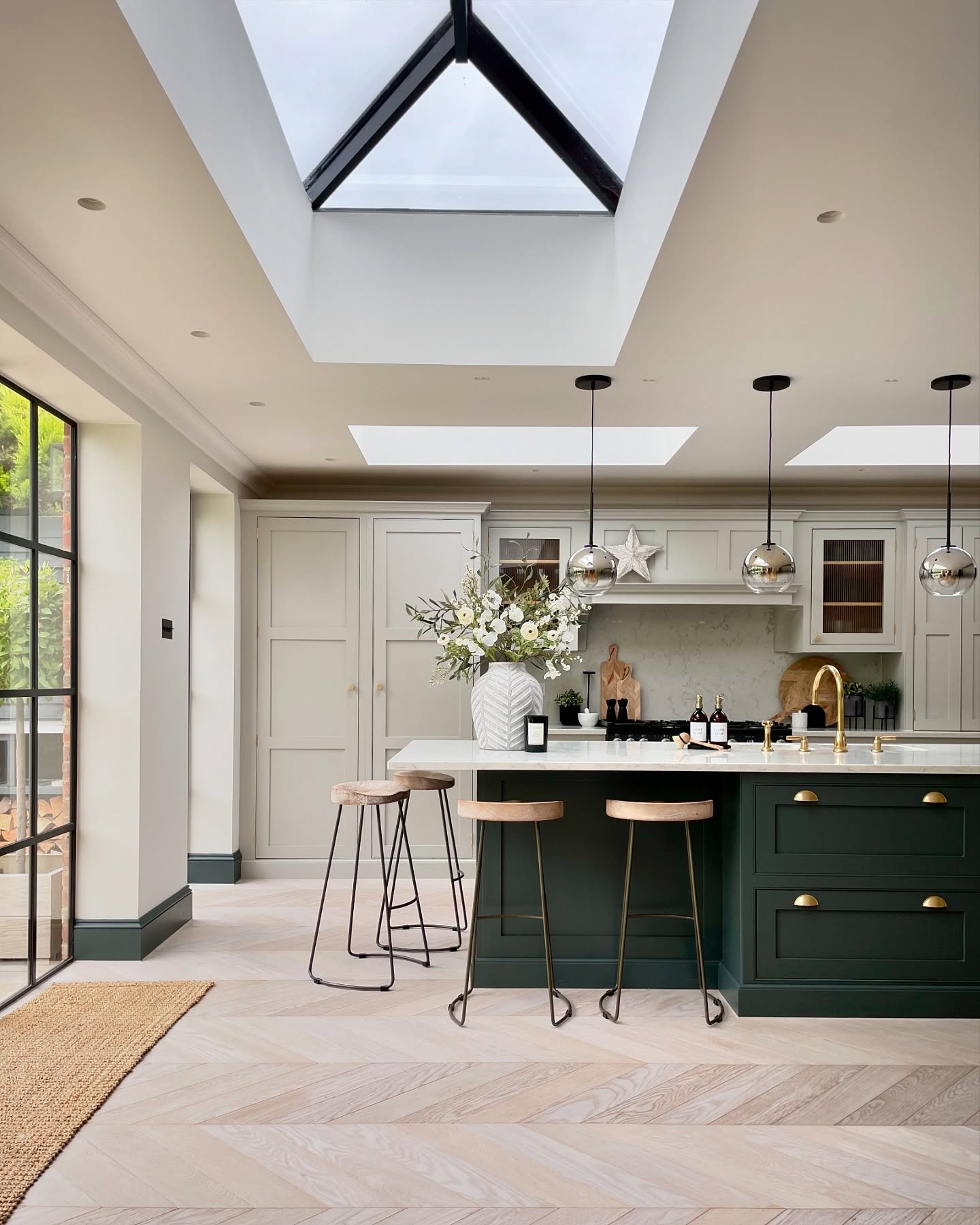 A dark green kitchen island with a white marble countertop and three wooden bar stools in a bright, modern kitchen with large skylights.
