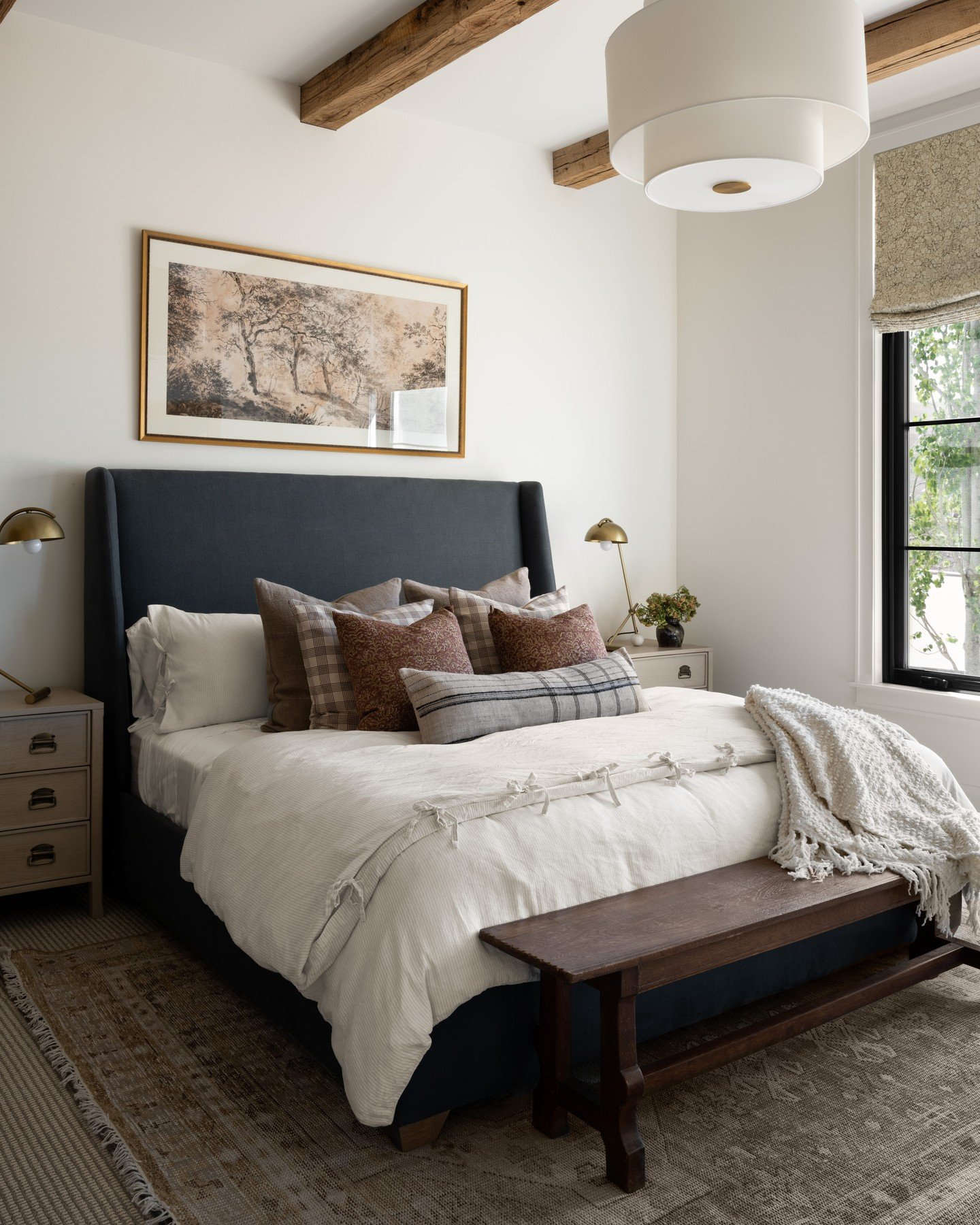 A sophisticated guest bedroom with a navy blue wingback bed, layered neutral bedding, and exposed wood beams. A rustic bench sits at the foot of the bed on a vintage-style rug, while a framed landscape painting hangs above the headboard.