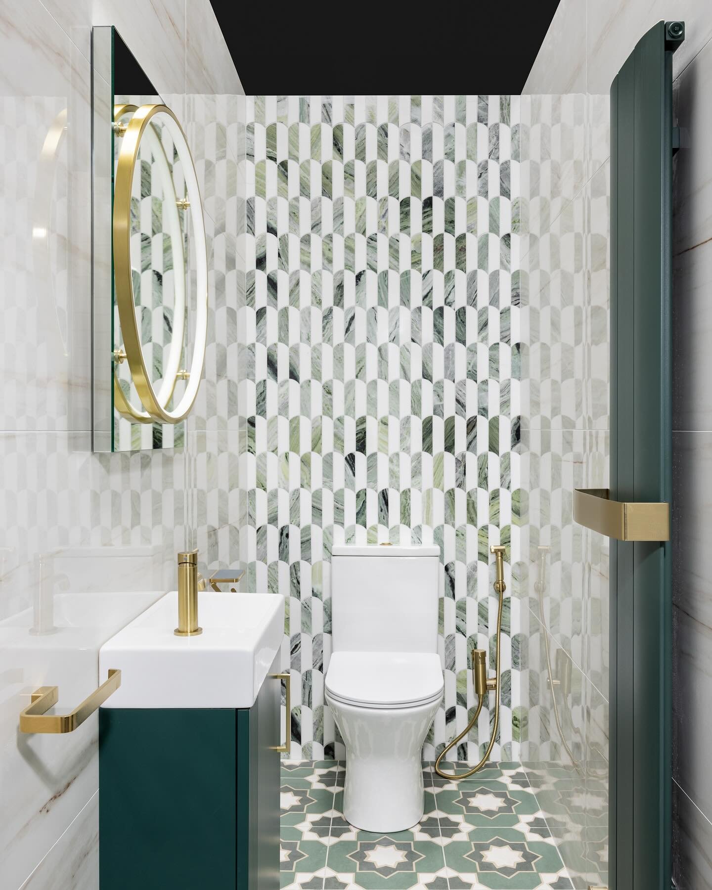 A small bathroom with a striking green and white geometric tile feature wall and a matching tiled floor. The space includes a dark green vanity with a vessel sink and an oval mirror with a gold frame.
