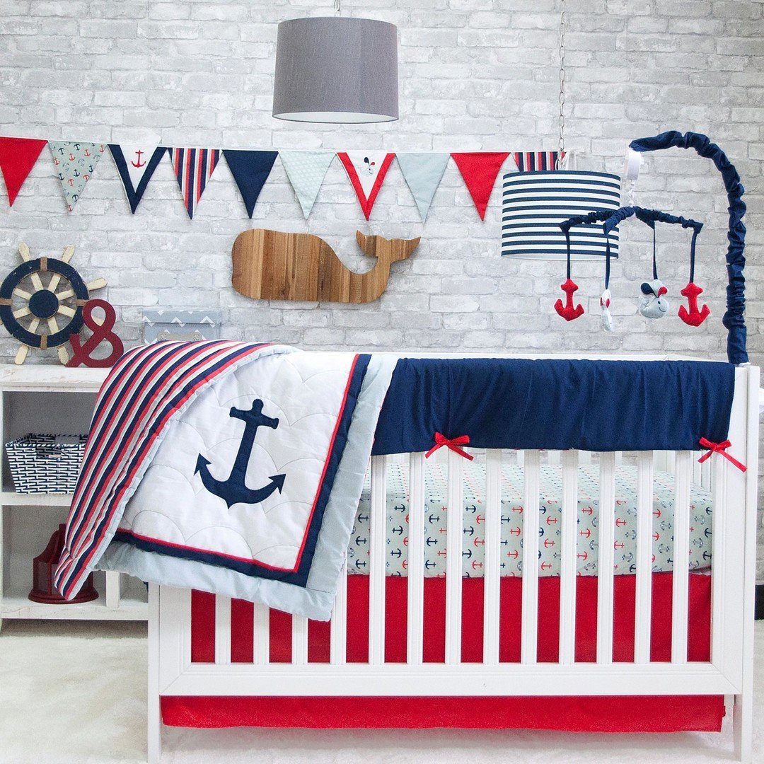 A nautical nursery with a white crib, red and navy blue anchor-themed bedding, a whale wall hanging, and a decorative banner.