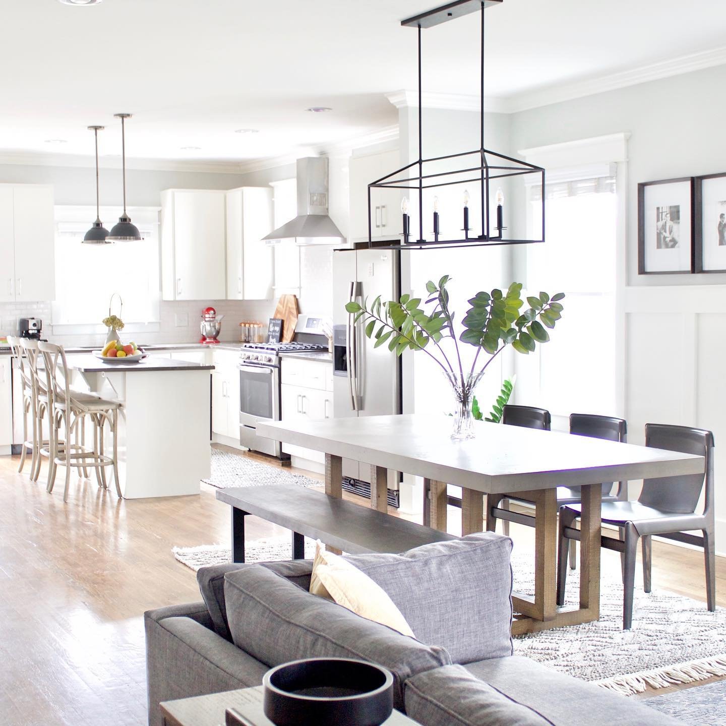 A bright, open-concept living, dining, and kitchen area with a modern farmhouse aesthetic. The living room features a grey sofa. A long wooden dining table with a bench and chairs separates the living area from the white kitchen with an island and stools.