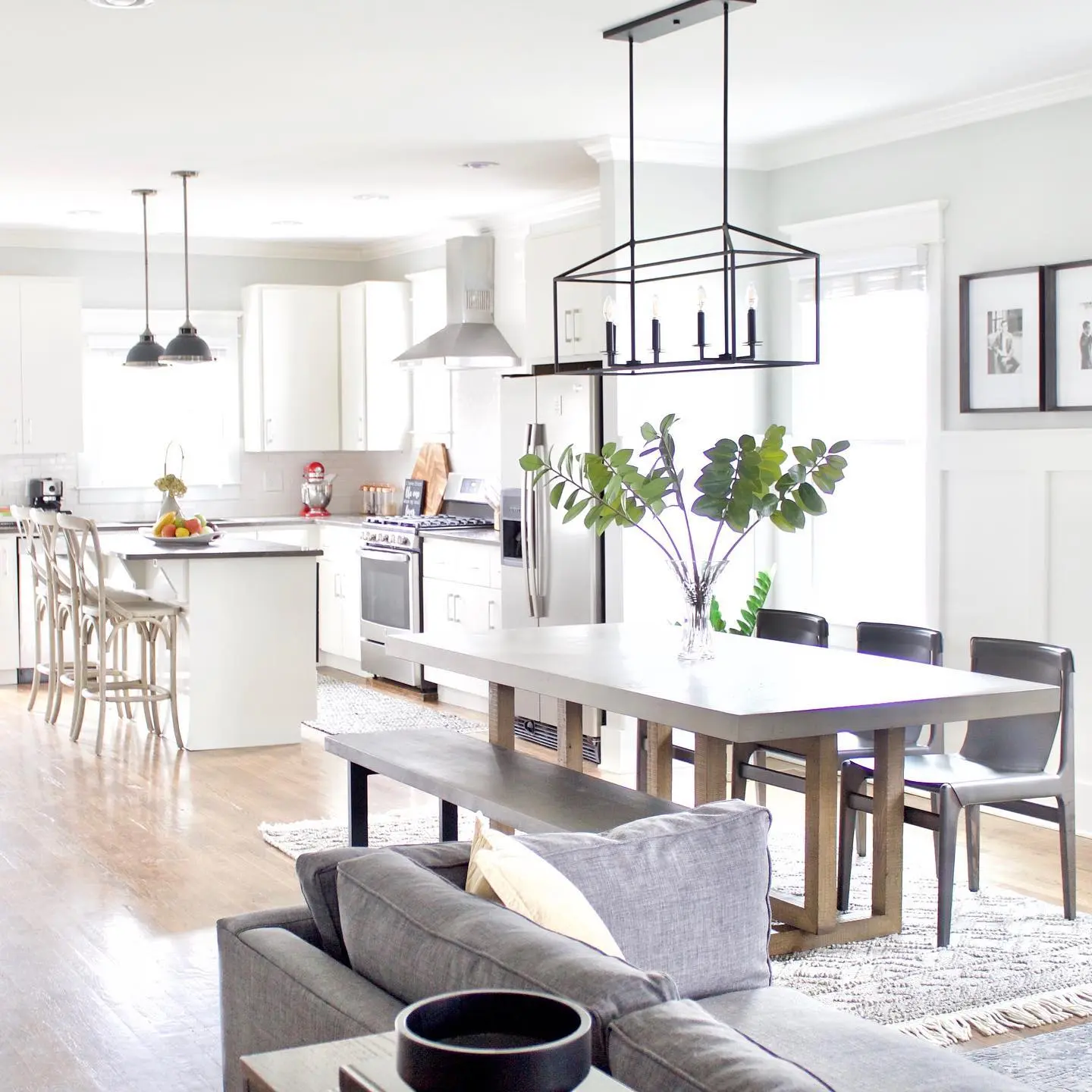 A bright, open-concept living, dining, and kitchen area with a modern farmhouse aesthetic. The living room features a grey sofa. A long wooden dining table with a bench and chairs separates the living area from the white kitchen with an island and stools.
