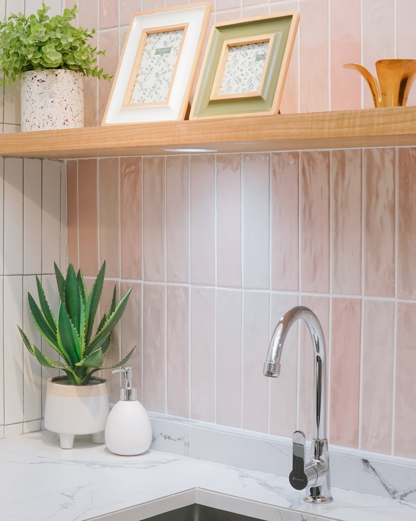 A close-up of a kitchen backsplash with glossy, vertical picket tiles in a soft blush pink color, paired with a white marble-effect countertop and wooden floating shelf.