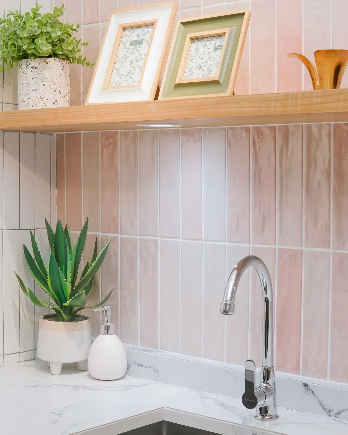 A close-up of a kitchen backsplash with glossy, vertical picket tiles in a soft blush pink color, paired with a white marble-effect countertop and wooden floating shelf.