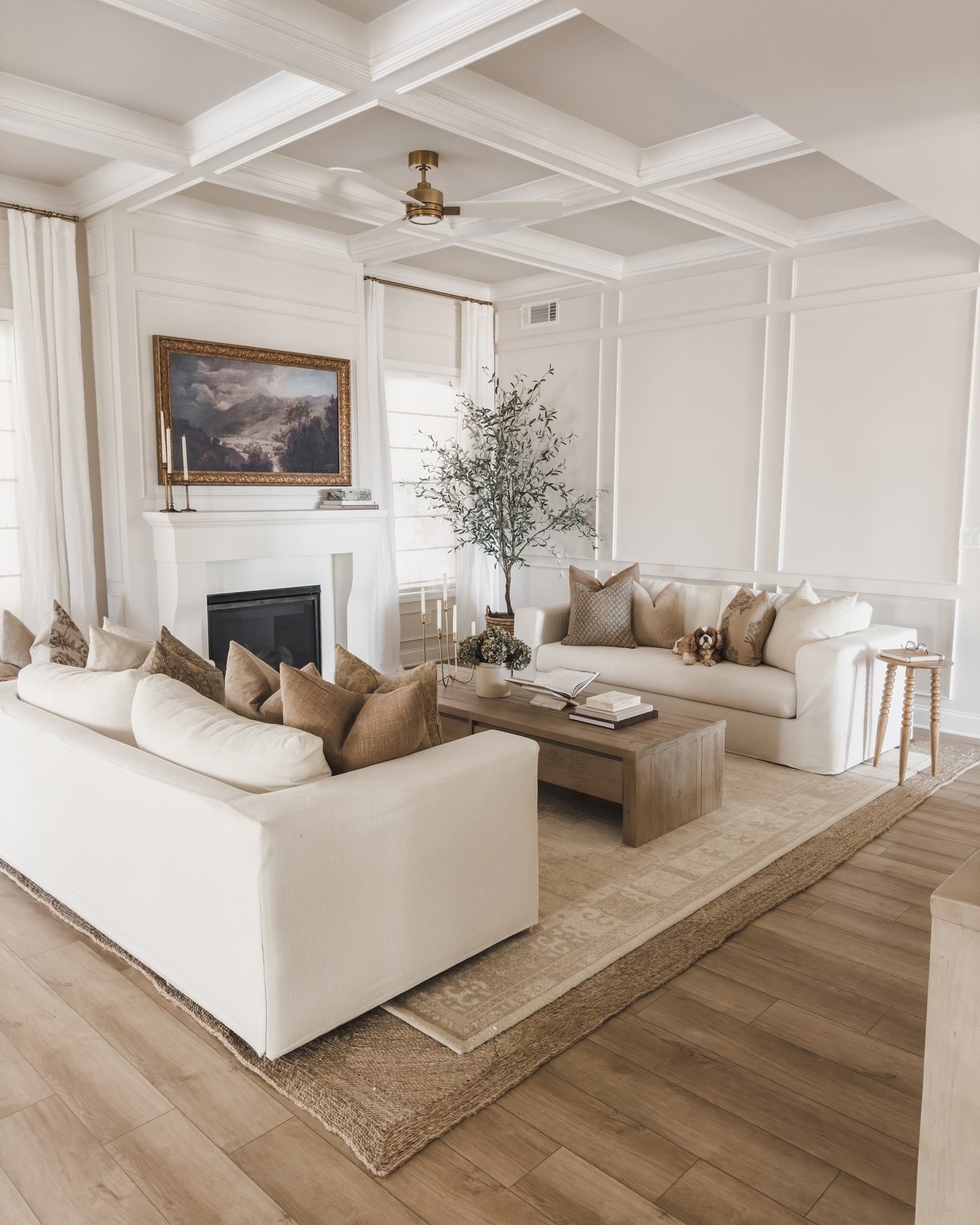 A bright and airy living room with two off-white sofas facing each other, a classic white fireplace, and a light wood coffee table on layered rugs.