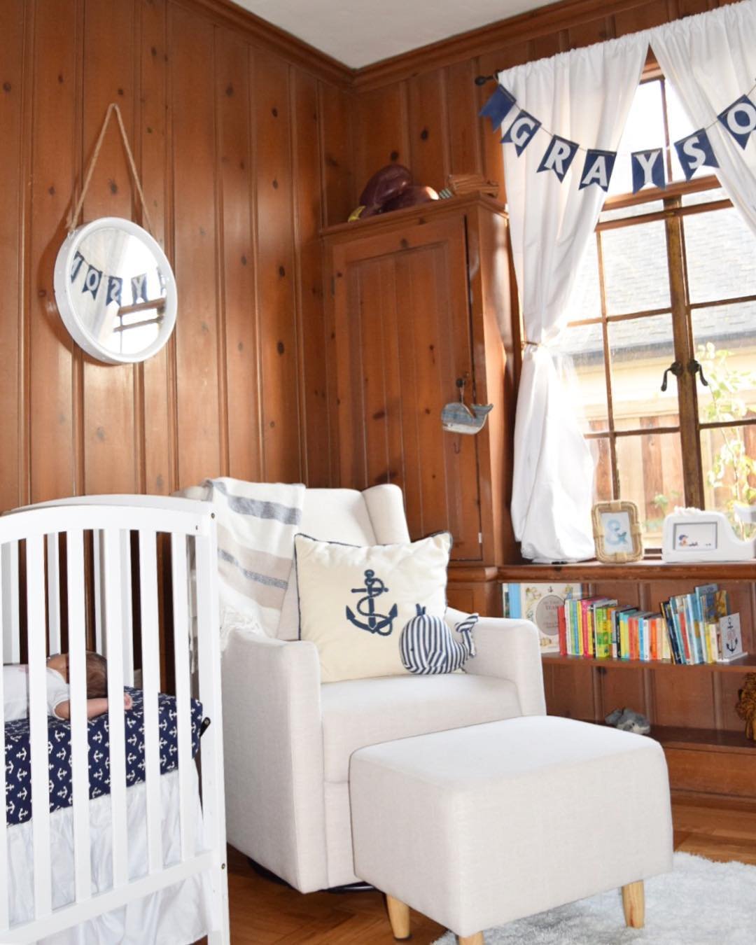 A cozy nursery with wood-paneled walls, a white crib, a glider chair, and personalized nautical-themed decorations.