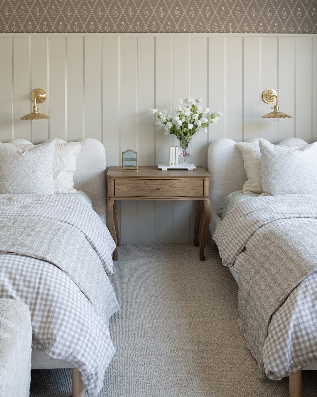 A cozy twin guest bedroom with two upholstered beds, a light wood nightstand between them, and a mix of vertical paneling and patterned wallpaper on the walls. Brass sconces are mounted on the wall above each bed.