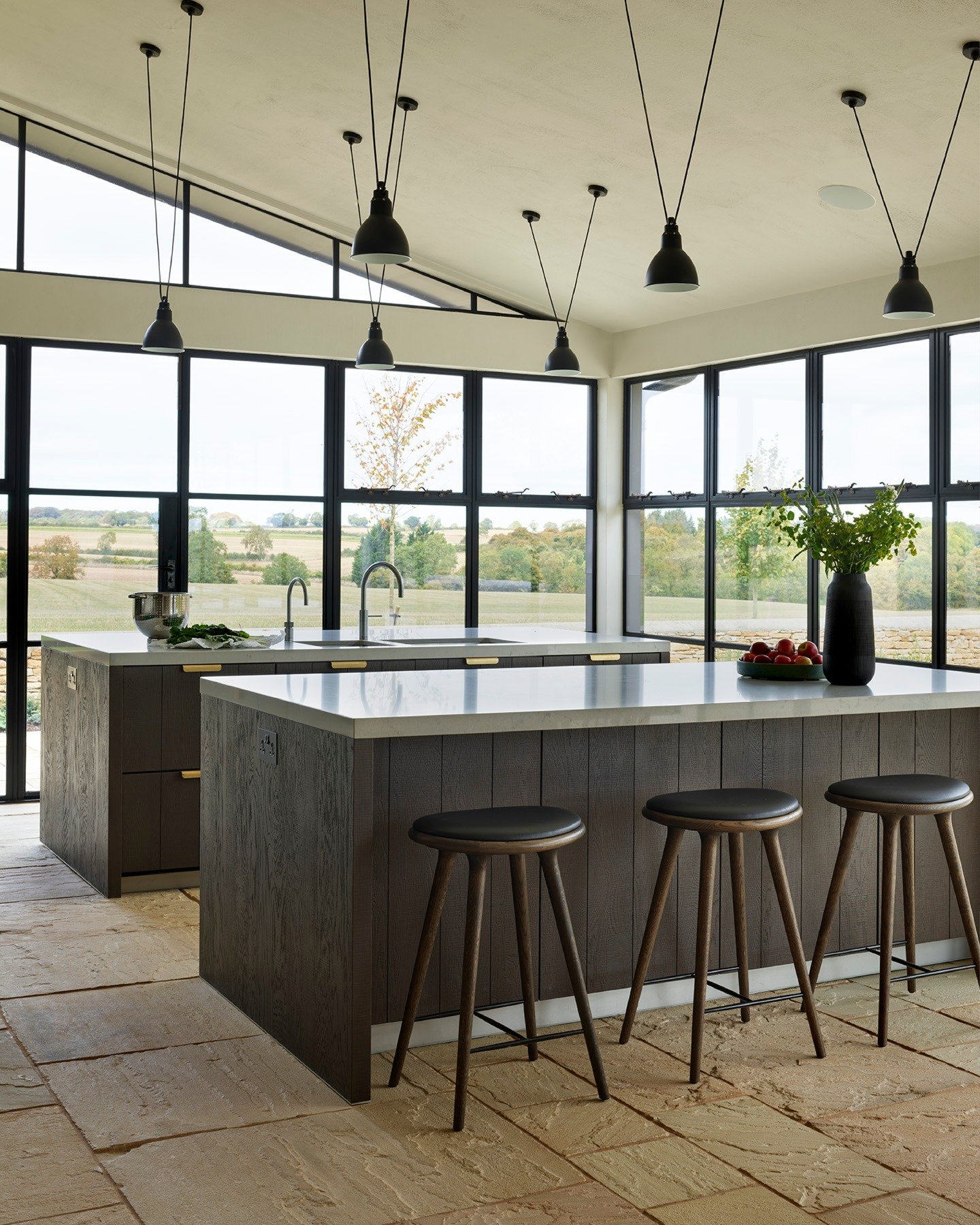 A spacious kitchen with two matching dark wood islands, white countertops, and black leather bar stools, surrounded by large windows.