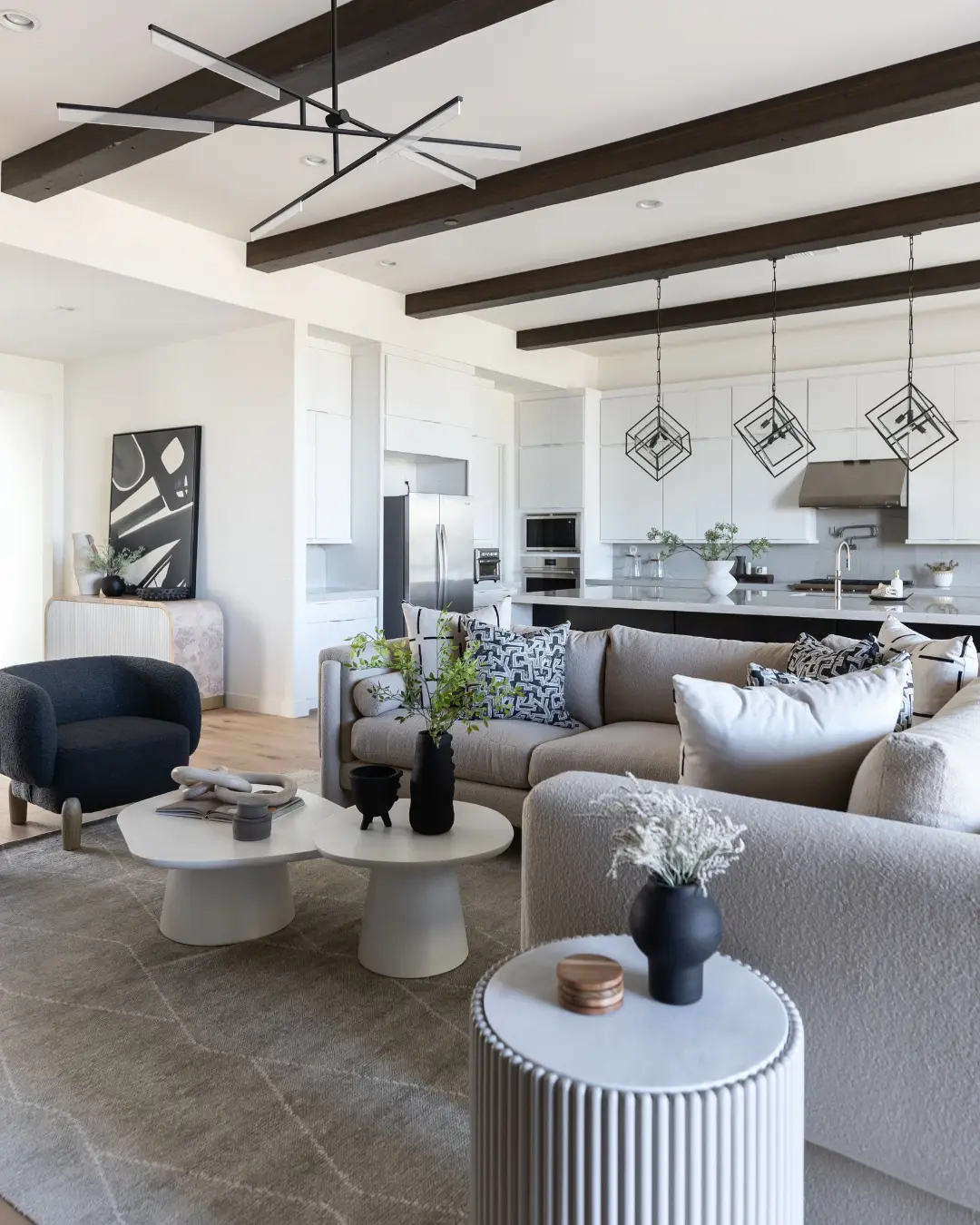 A modern, open-concept living and kitchen area with dark wood ceiling beams. The living space features two light-toned sofas, two white oval coffee tables, and a dark grey armchair. The kitchen has white cabinetry, a large island, and geometric pendant lights.