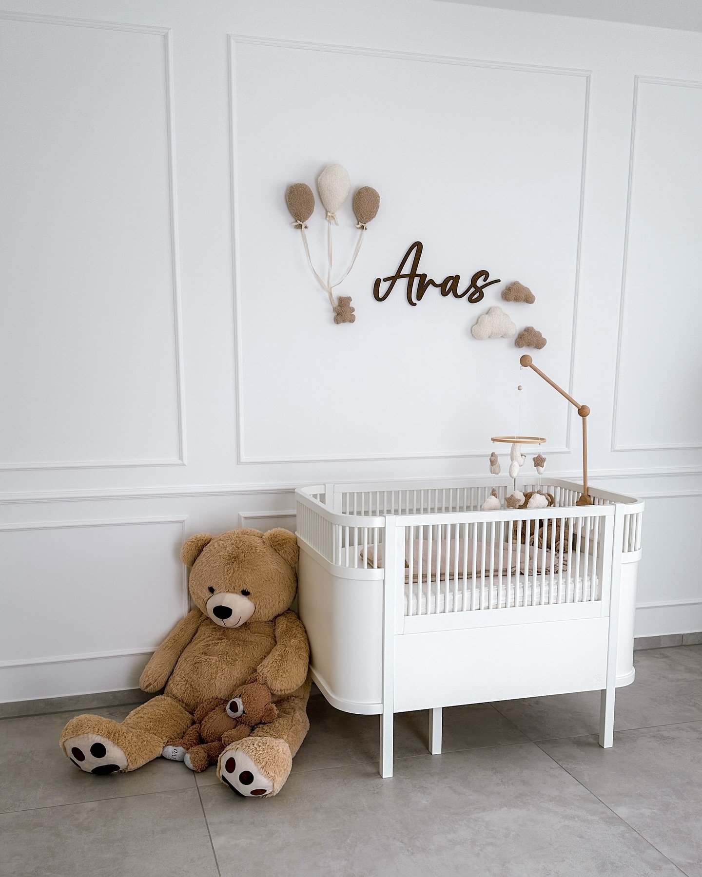 A minimalist white nursery room with a modern crib, a custom name sign on the wall, and two teddy bears on the floor.