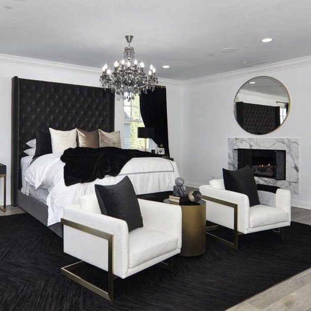 A glamorous black and white bedroom with a tall, tufted black headboard, a crystal chandelier, two white armchairs with gold frames, and a marble fireplace.