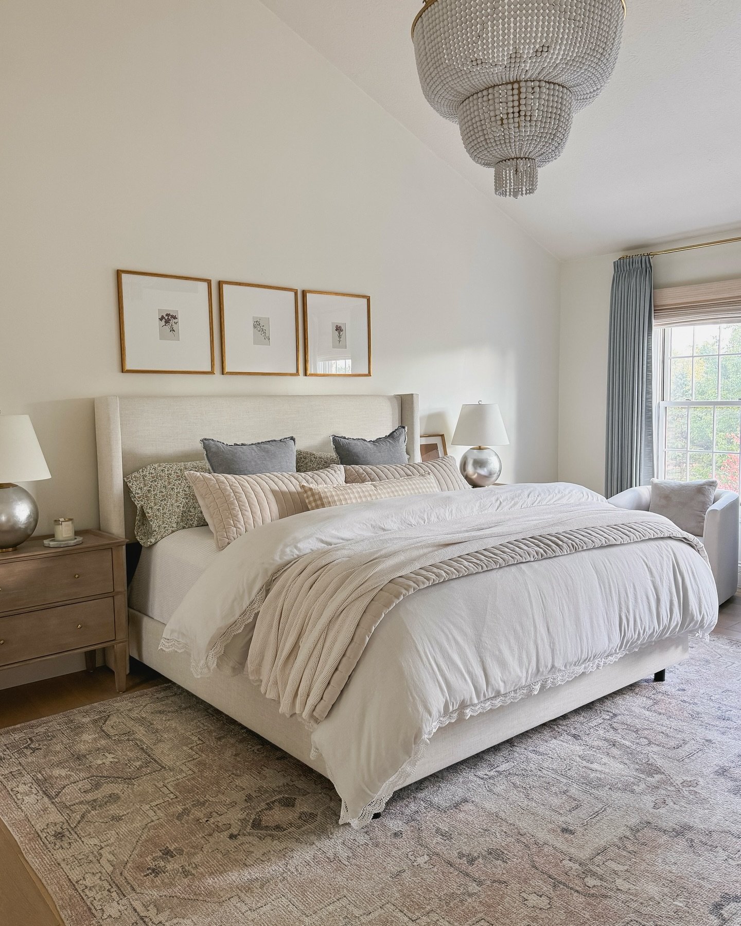A large area rug with a faded pattern in shades of cream, gray, and blush is placed under a large bed with a beige upholstered headboard.
