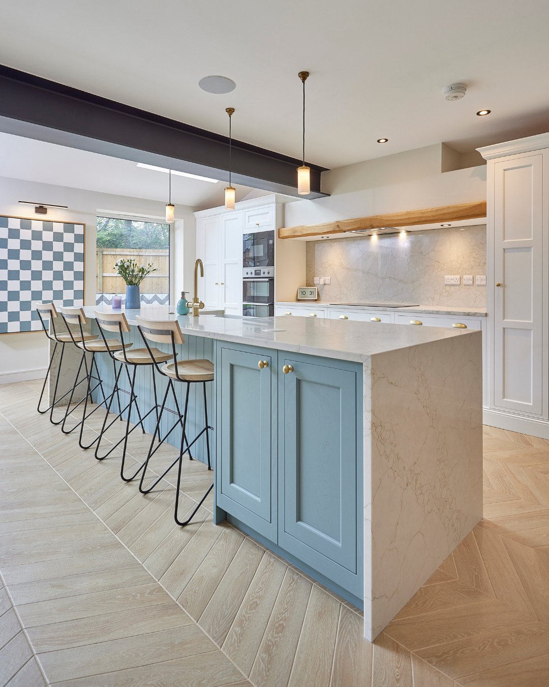 A long, light blue kitchen island with a marble waterfall countertop and four wooden bar stools in a bright, contemporary kitchen.