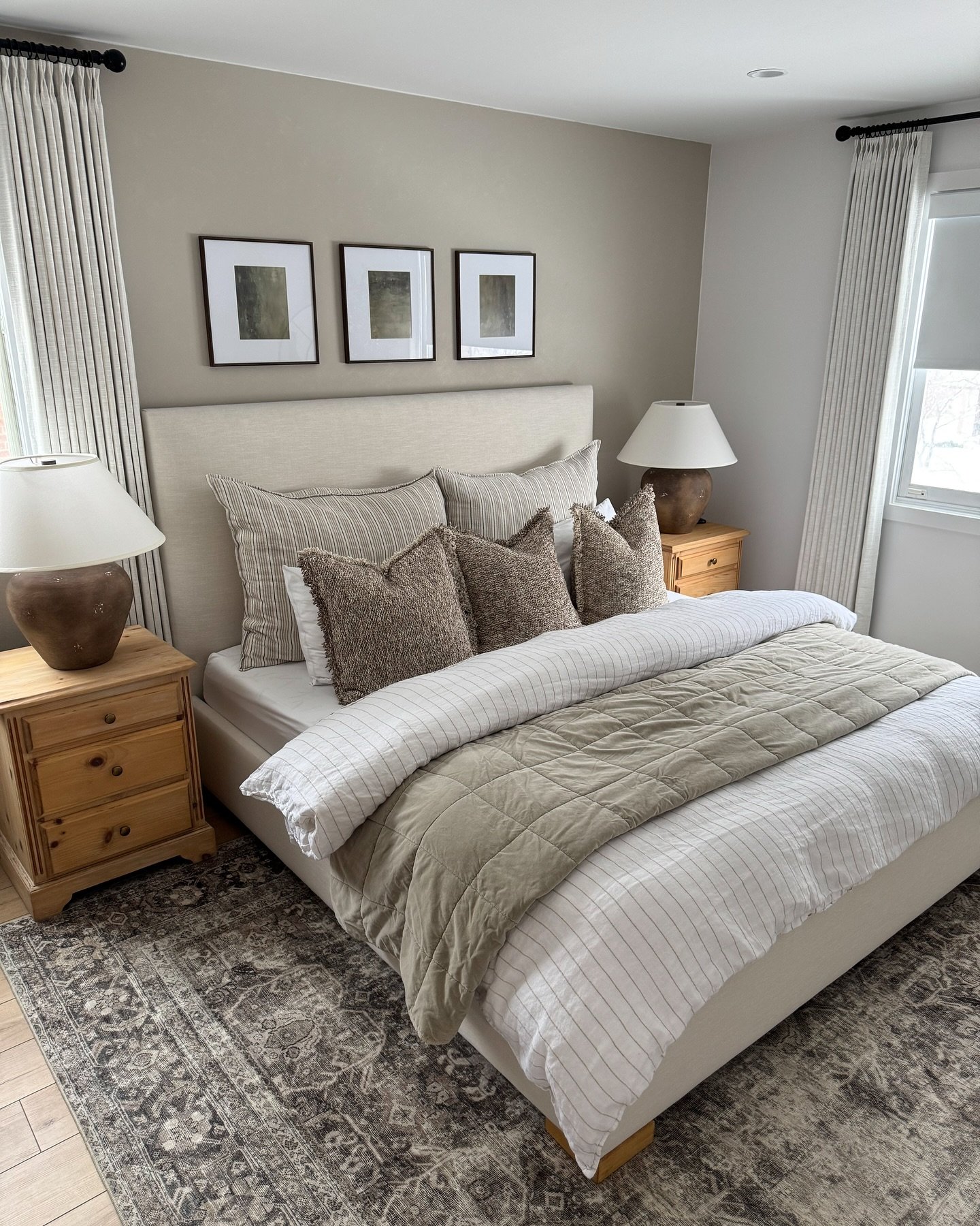 A neutral and inviting guest bedroom with an upholstered bed, striped and textured bedding, and rustic wood nightstands. The room features a gallery wall of three black-framed prints and a patterned area rug.